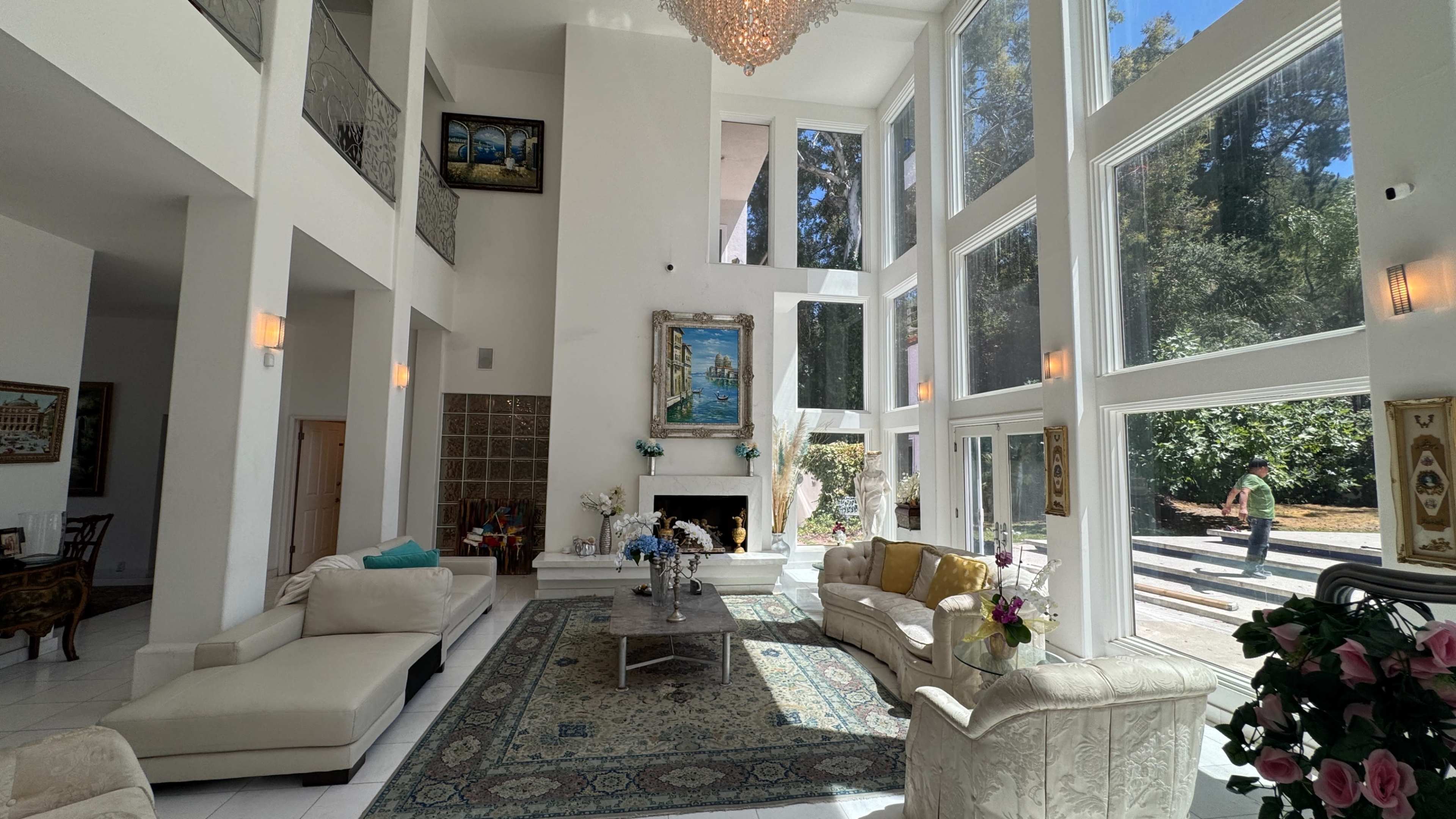 The interior of a spacious living room features large windows, a chandelier, and elegant furniture arranged around a central coffee table.