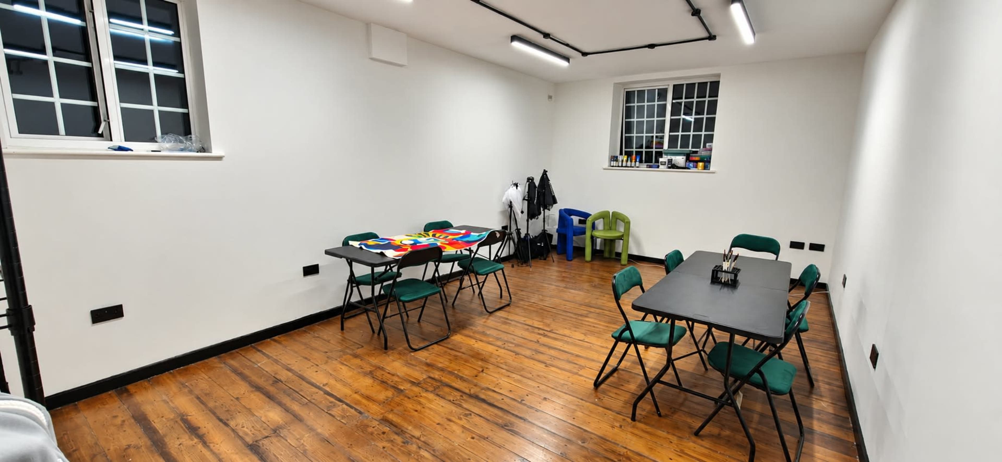 The image shows a vacant room with wooden floors, two folding tables surrounded by green chairs, and a few items hanging on the wall.