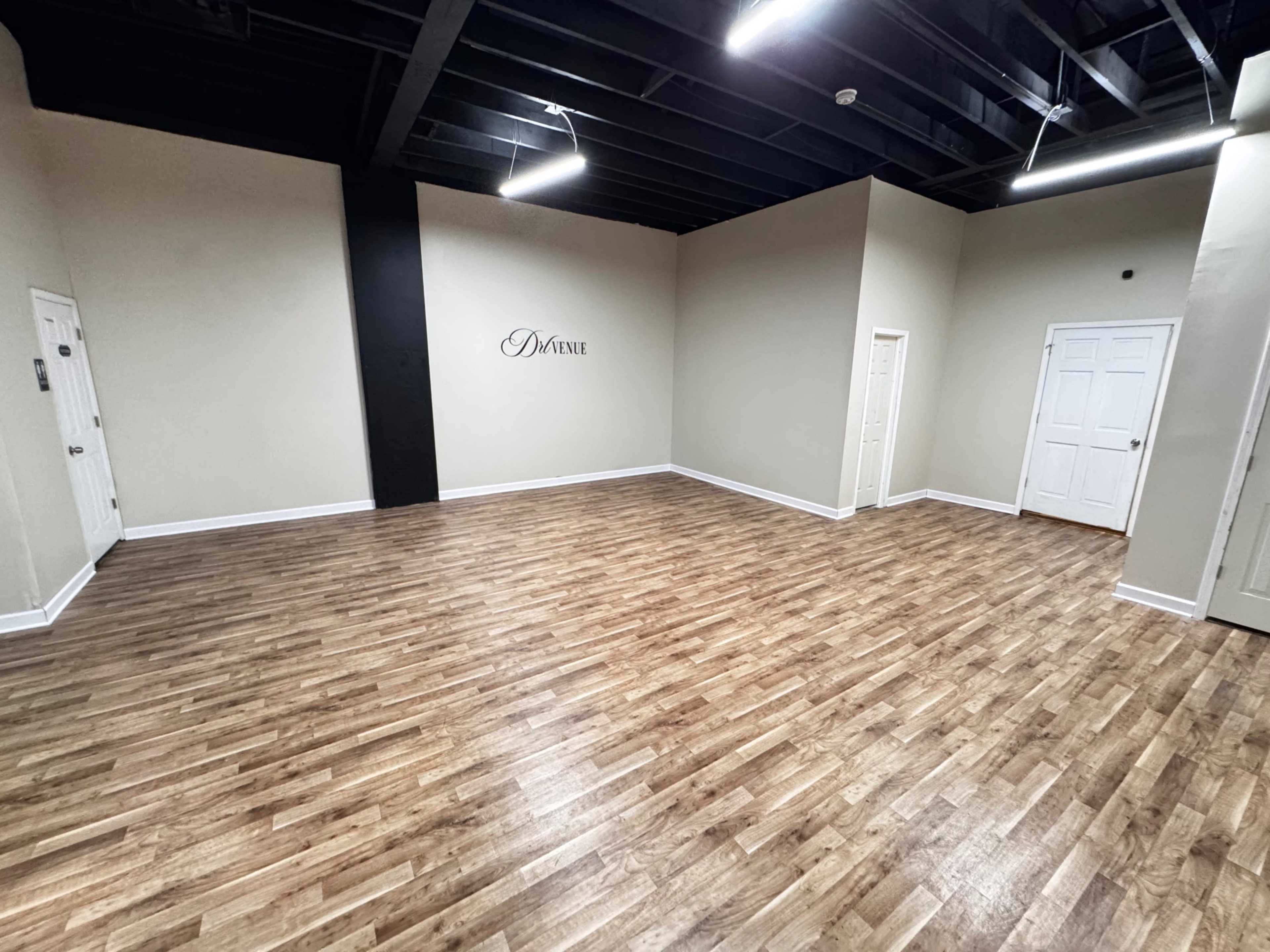 The image shows a spacious, empty room with wooden flooring, beige walls, and a black accent wall featuring the word "Divine."
