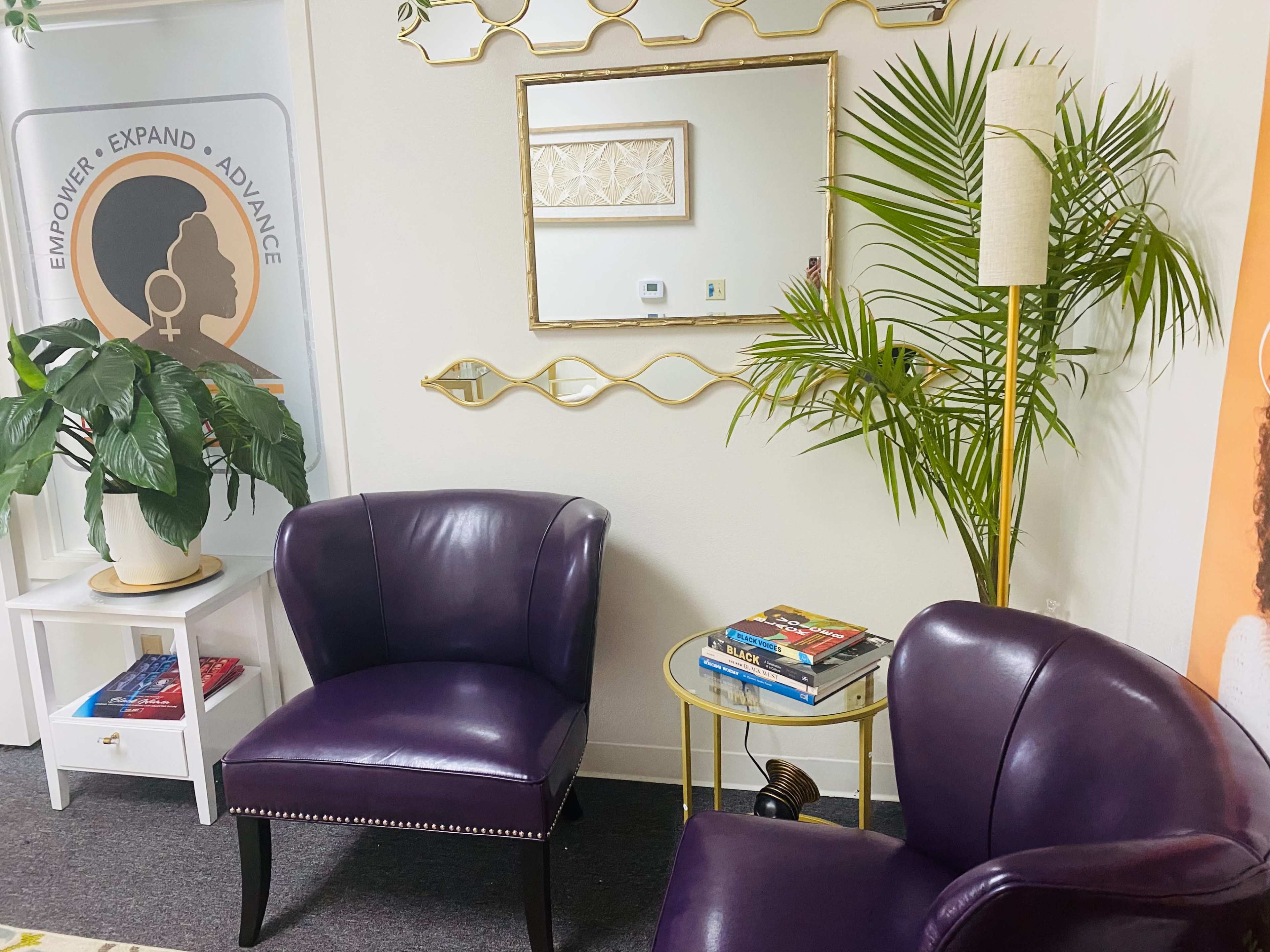 The image shows a cozy seating area with two purple armchairs, a small white table, a decorative mirror on the wall, a potted plant, and several books on the floor.