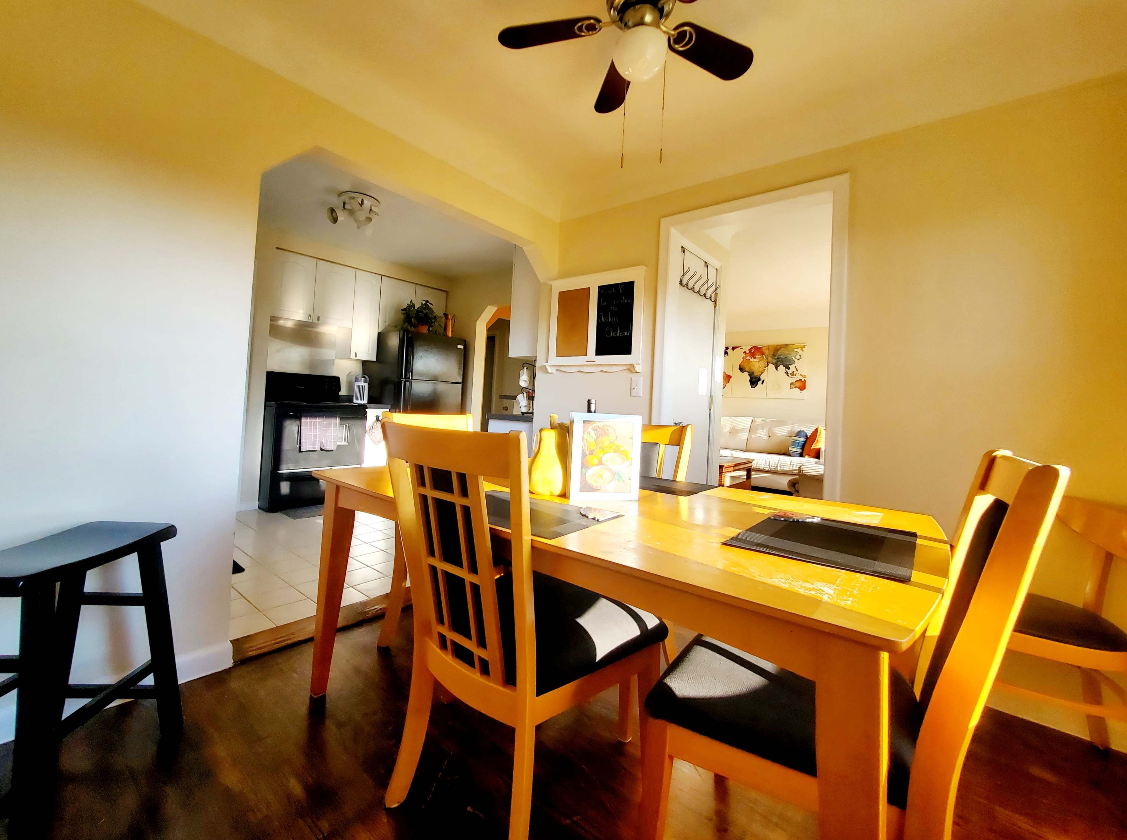 A dining area features a wooden table surrounded by chairs, with a doorway leading to a kitchen and another room in the background.