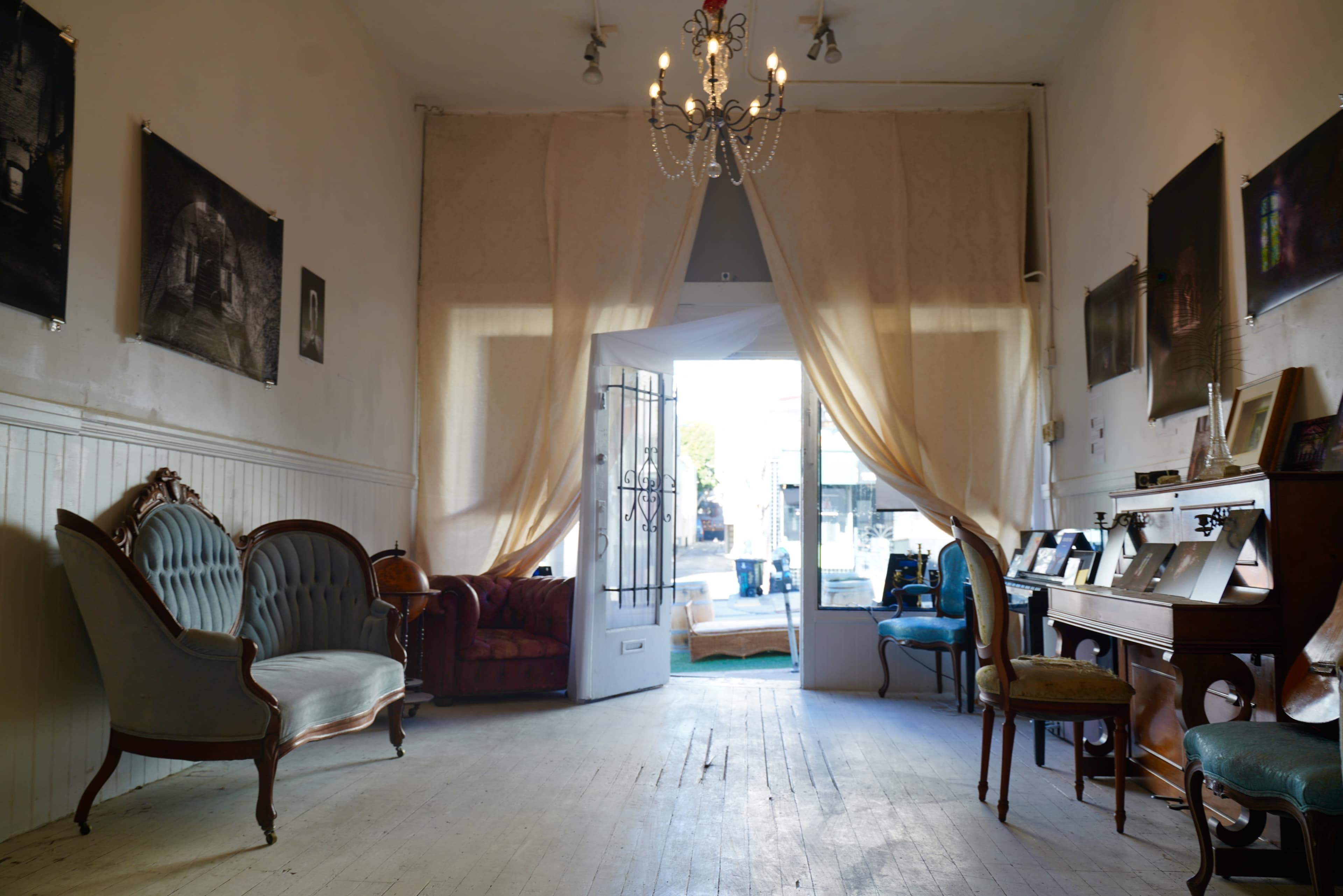 A lightly furnished room with two vintage sofas, a piano, and an open door leading outside, adorned with soft drapes and framed photographs on the walls.