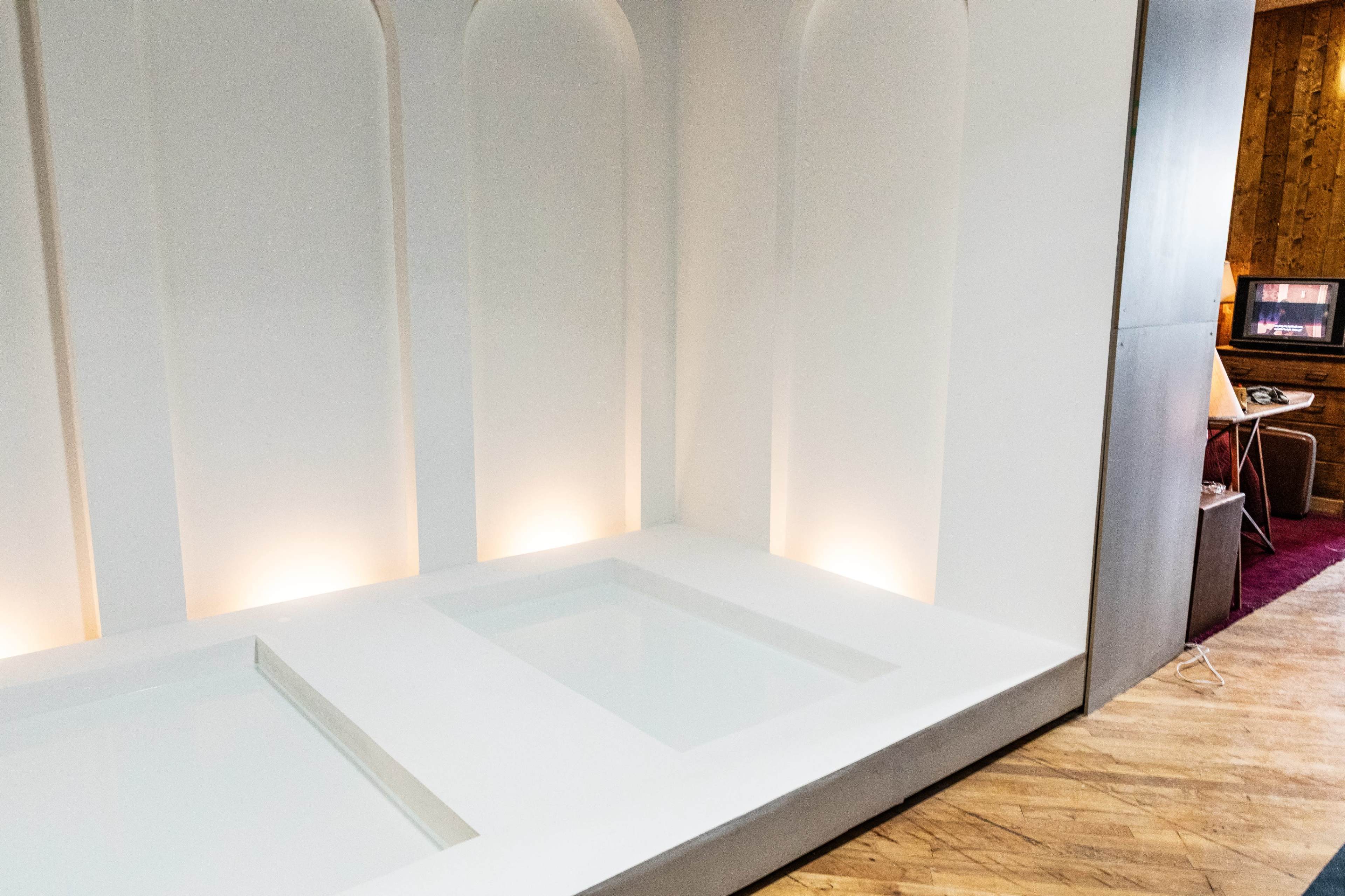 The image shows a minimalist room featuring a white, two-sectioned bathing area with recessed lighting along the walls.