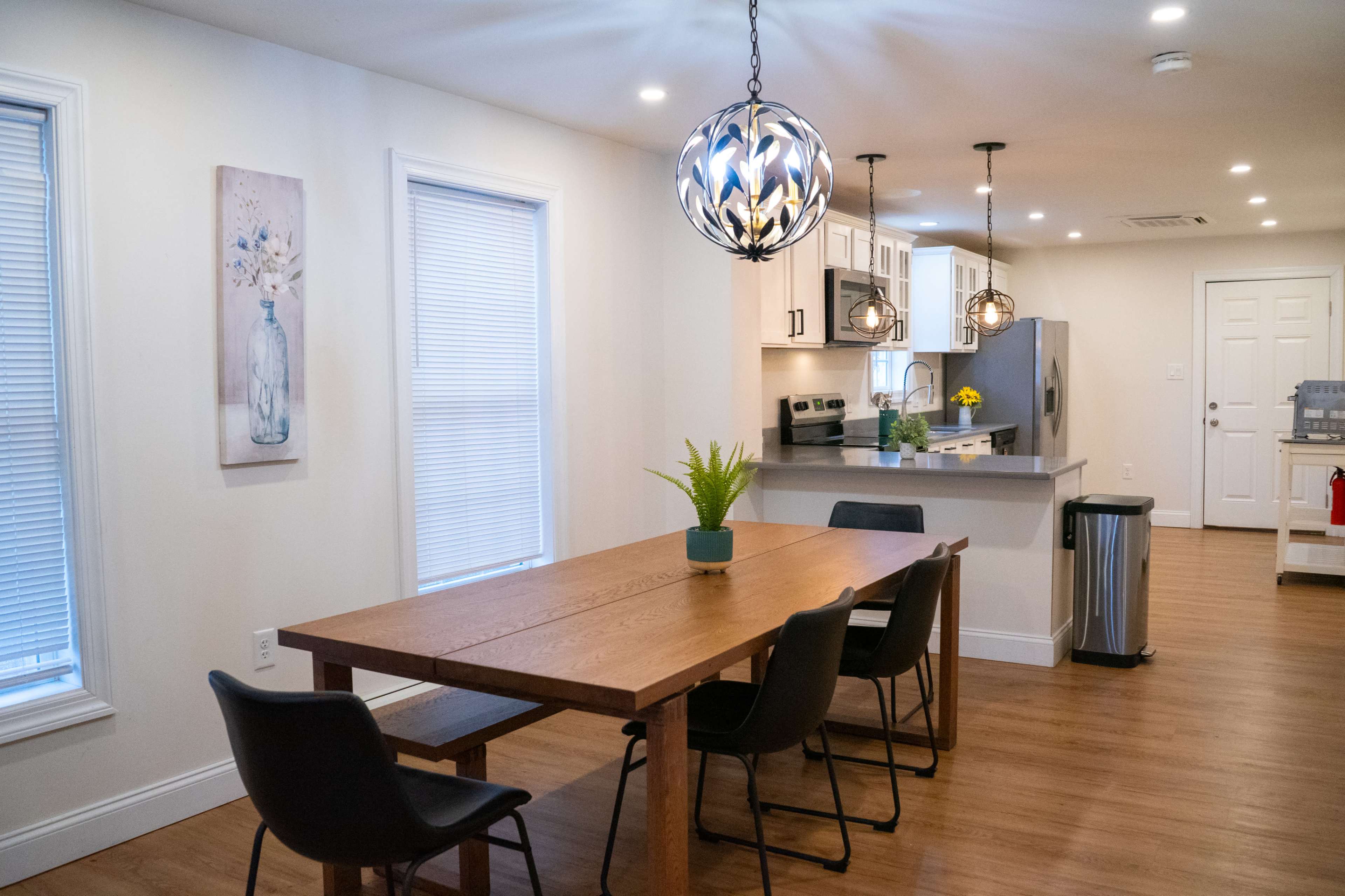 A modern dining area with a wooden table, four black chairs, and a kitchen visible in the background, featuring light cabinets and pendant lighting.