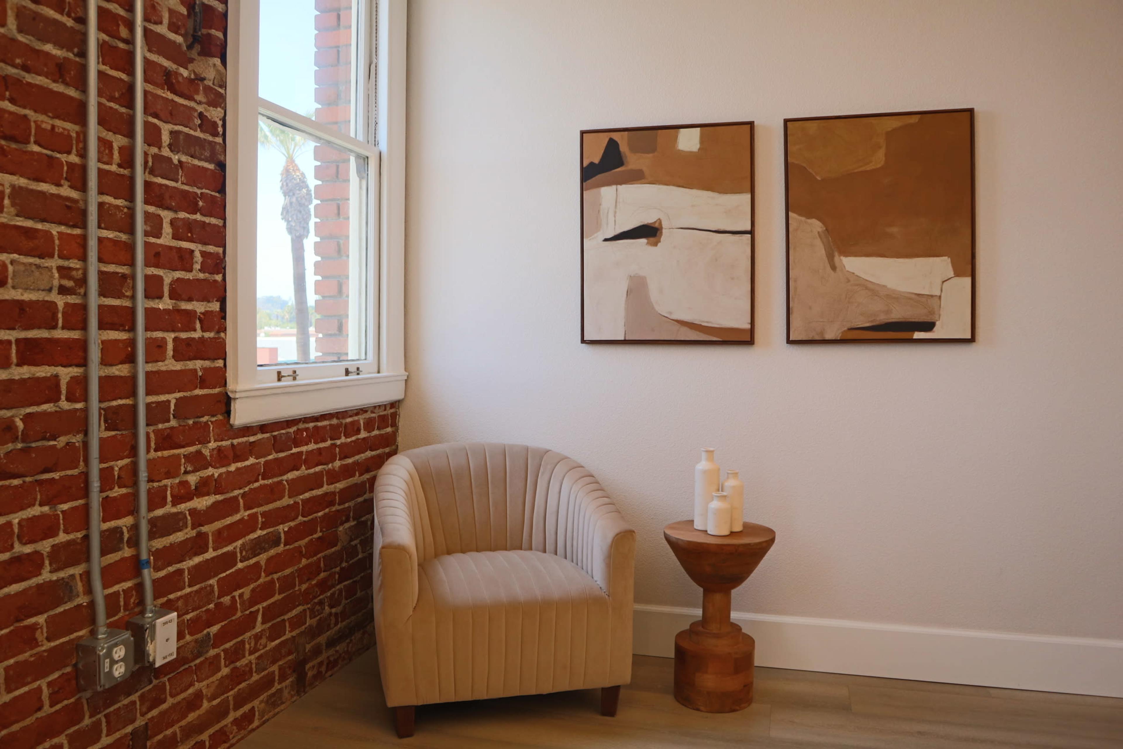 A corner of a room featuring a plush, light-colored armchair next to a round wooden side table with three white containers, against a backdrop of exposed red brick walls and two abstract paintings.