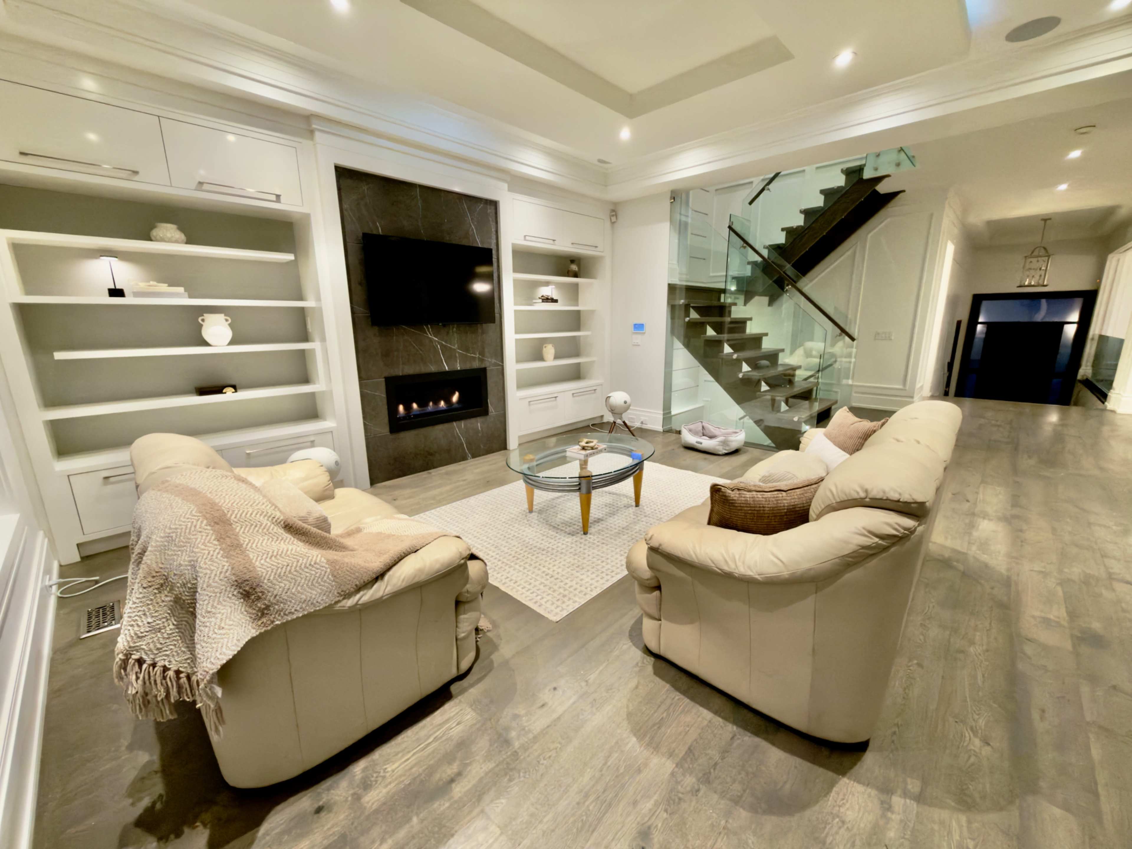The image shows a modern living room with two beige leather sofas facing a glass coffee table, situated under a sleek wall-mounted TV and a decorative fireplace.
