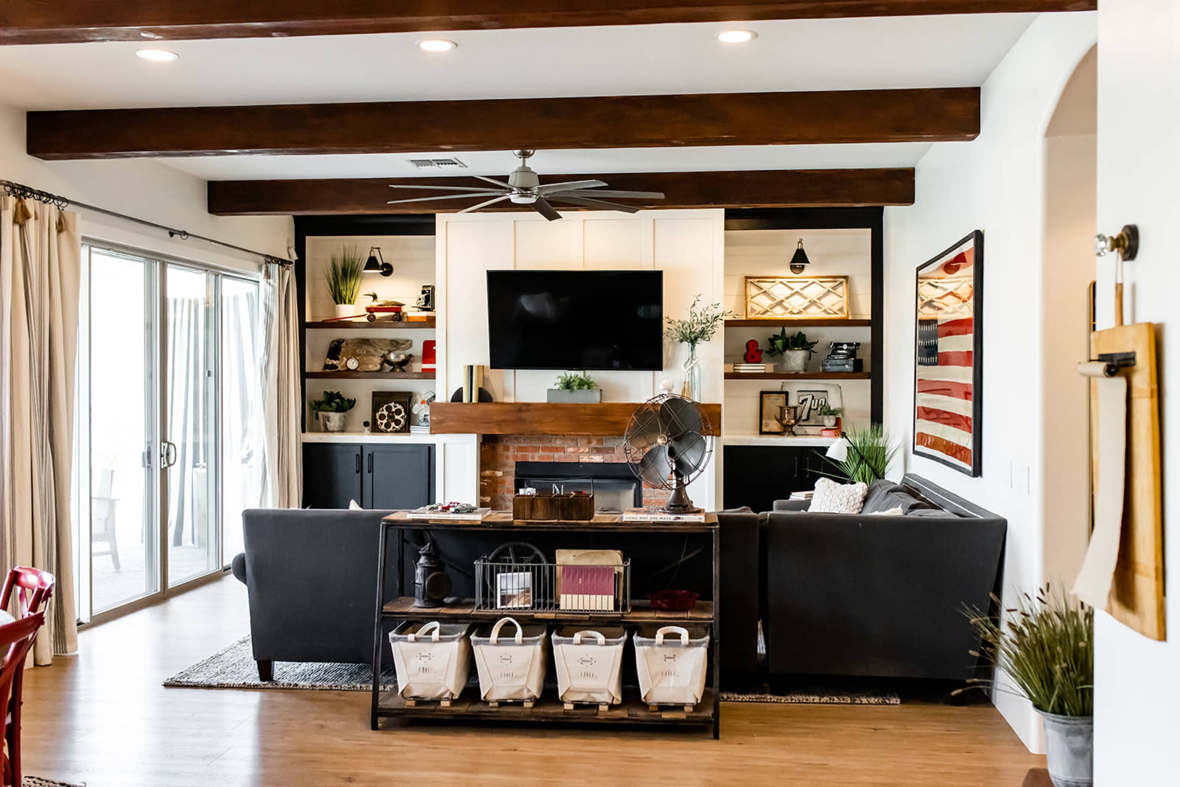 The living room features a black sofa, a wooden coffee table with storage bins, and a wall-mounted TV above a fireplace, surrounded by built-in shelves with decorative items.