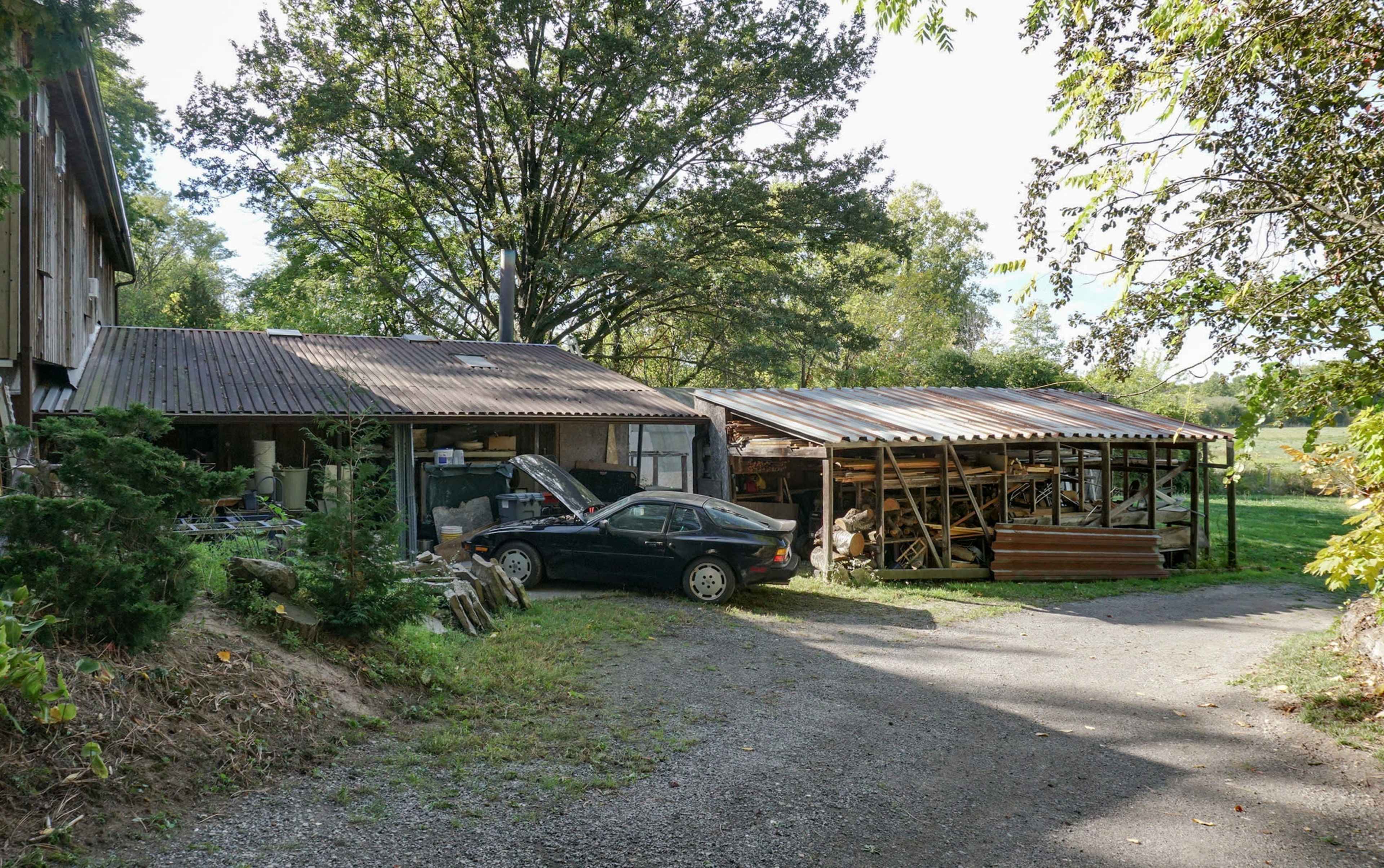 The image shows a gravel driveway leading to two structures, one with a car parked in front and the other open, displaying piles of wood and equipment.
