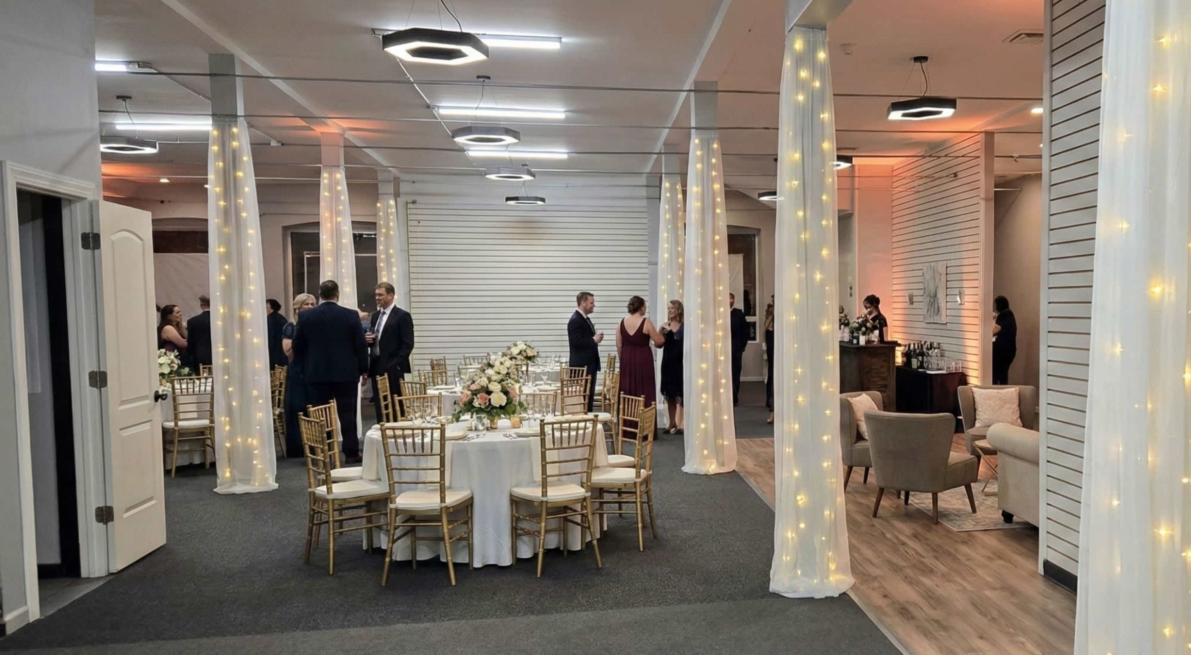 The image shows a spacious event venue with tables set for dining, elegantly decorated with flowers, and guests mingling in formal attire.