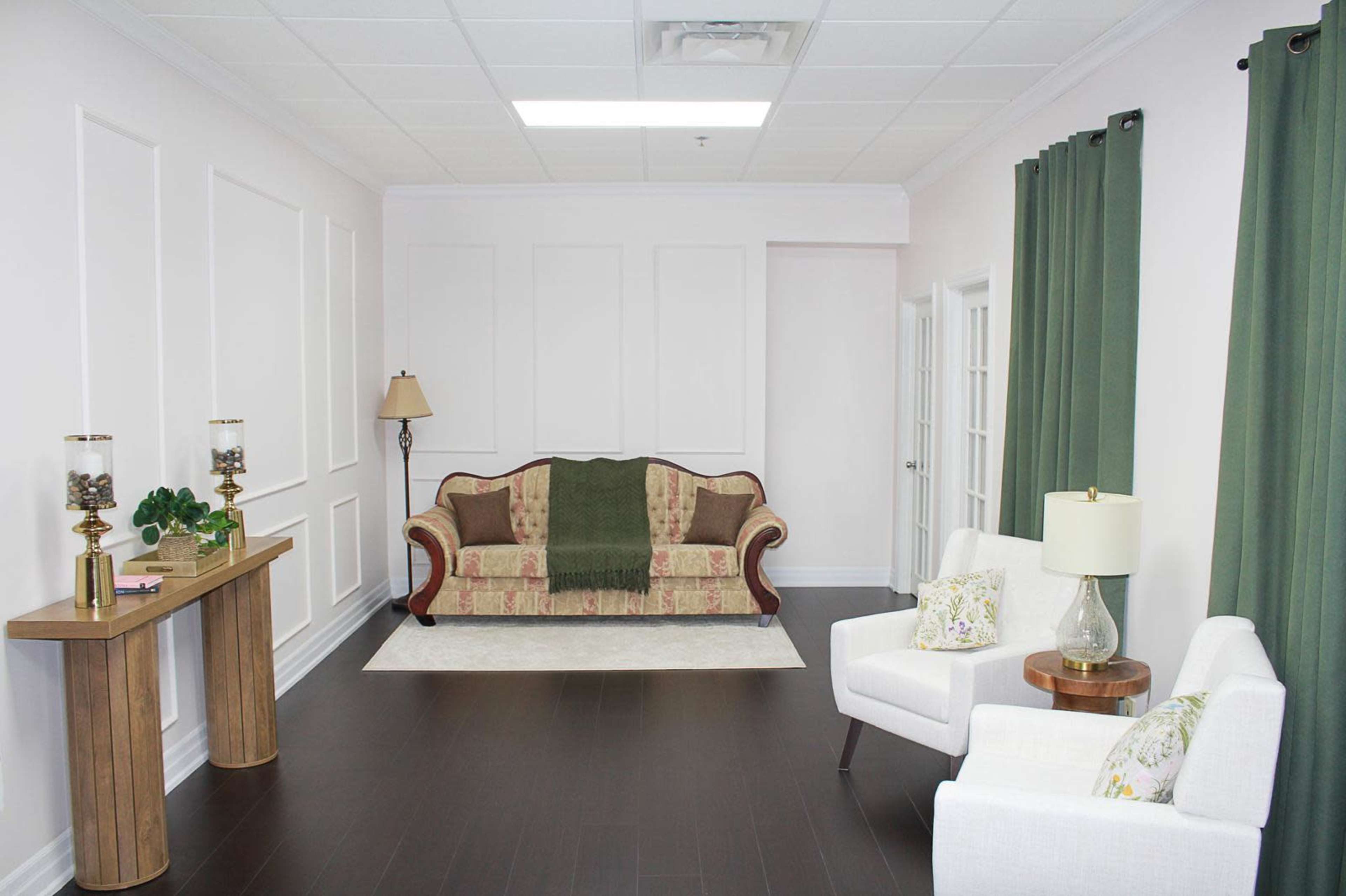 The image shows a well-lit room featuring a patterned sofa, a pair of white armchairs, a decorative table, and green curtains.
