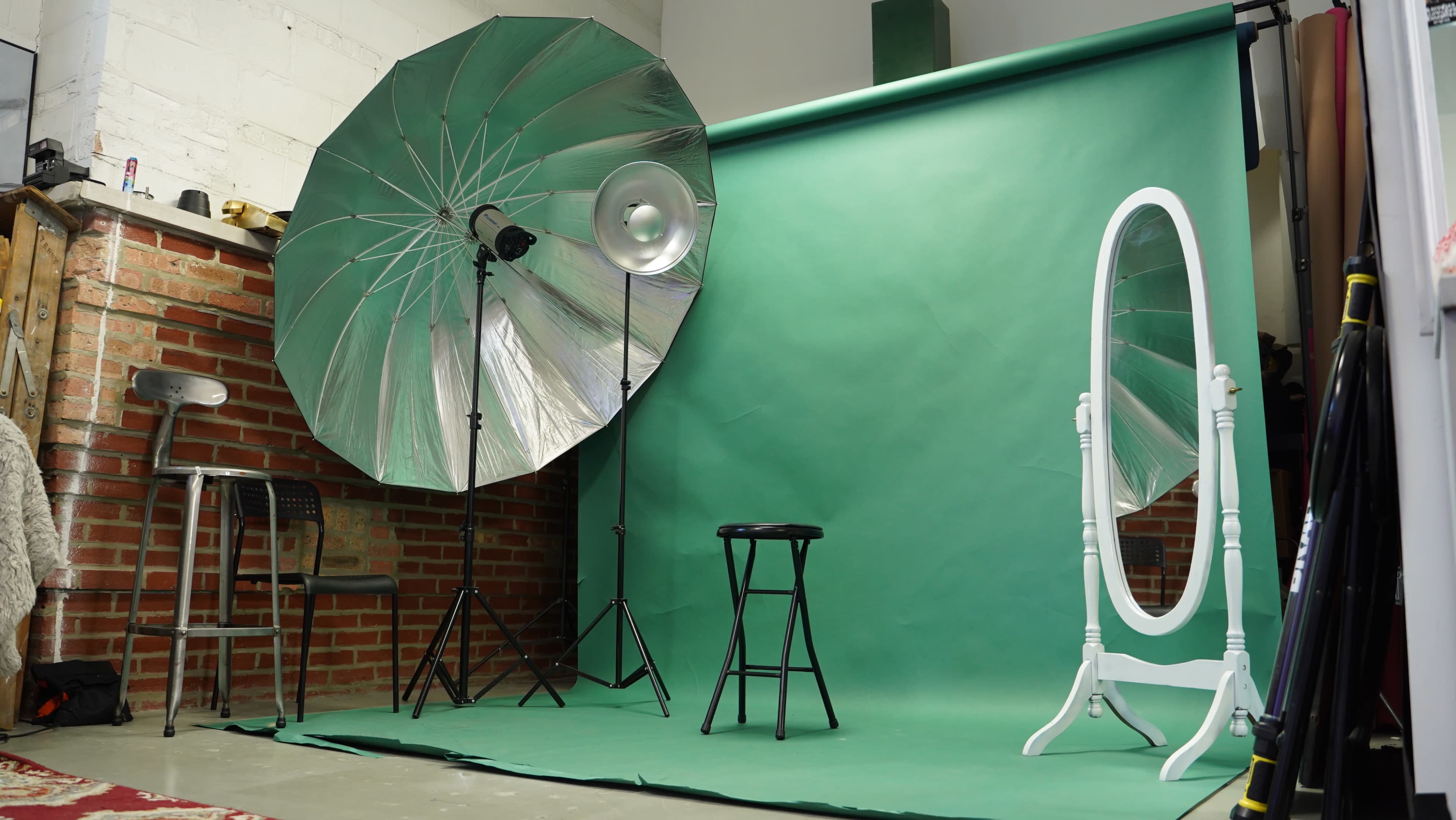 A photography studio equipped with a large umbrella light, a green backdrop, a stool, and a mirror.