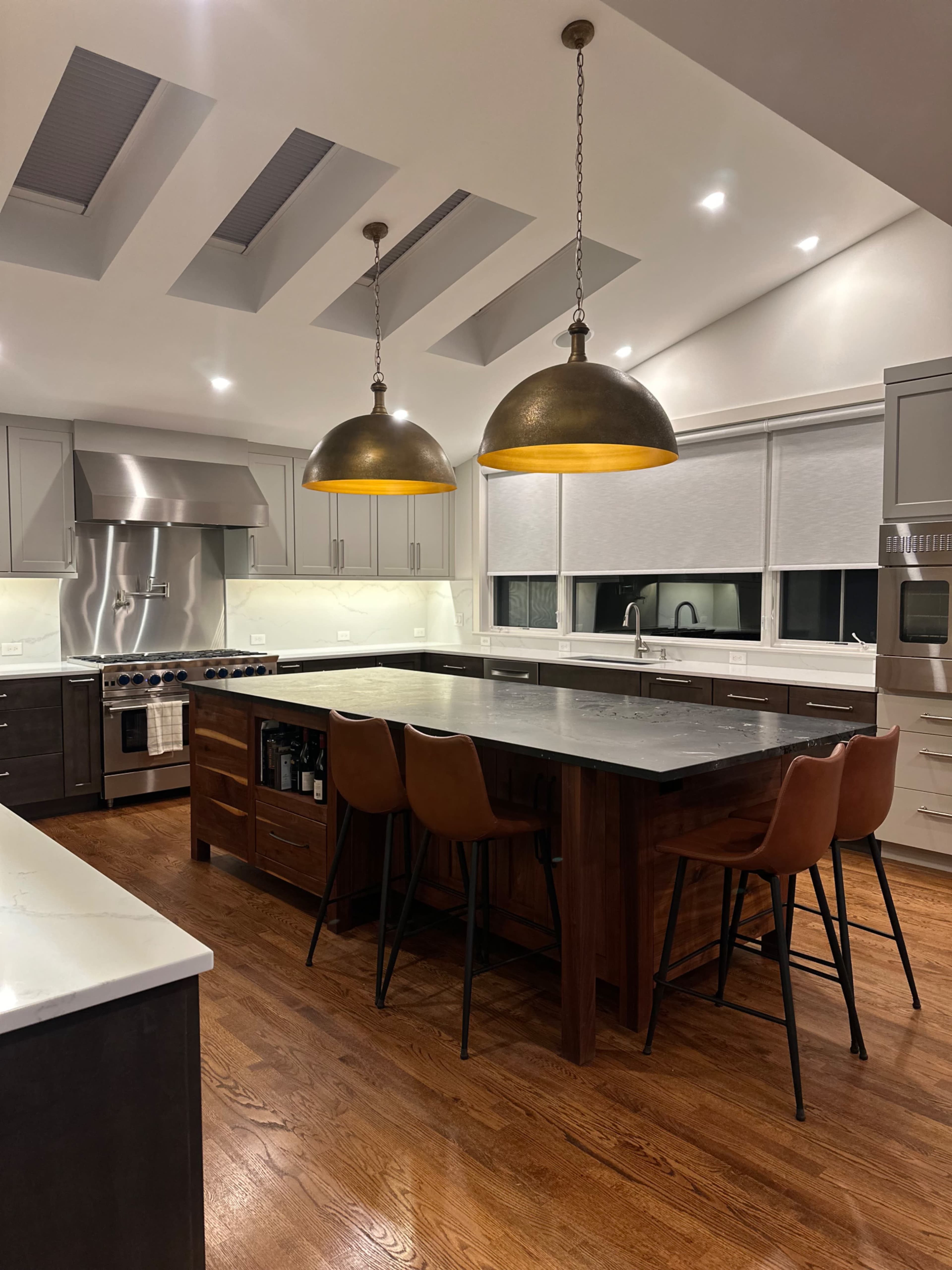 The image shows a modern kitchen featuring a large central island with four brown stools, pendant lights above, stainless steel appliances, and white cabinetry.