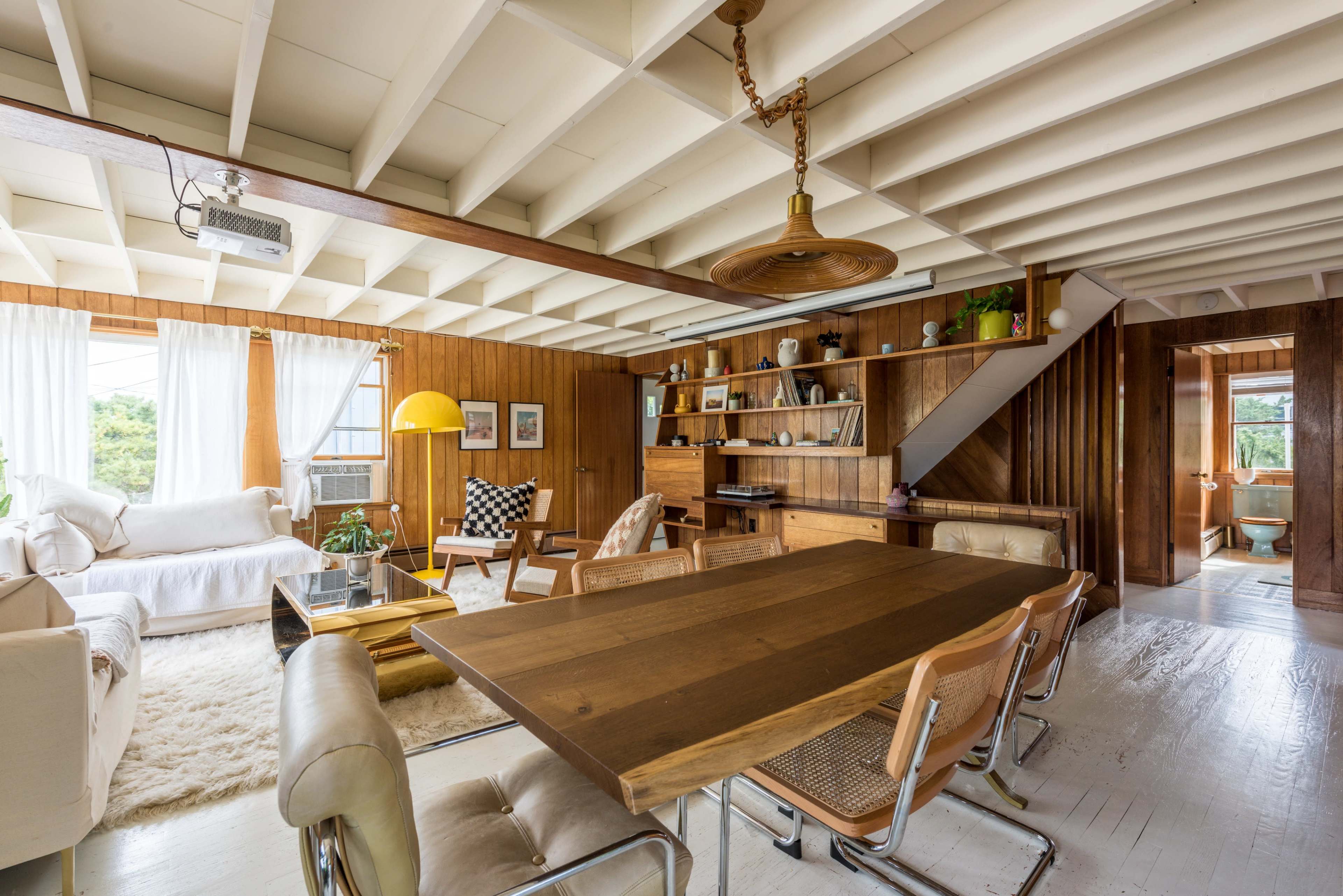 A spacious living and dining area featuring a wooden table, chairs, a light-colored sofa, and a large window with curtains.