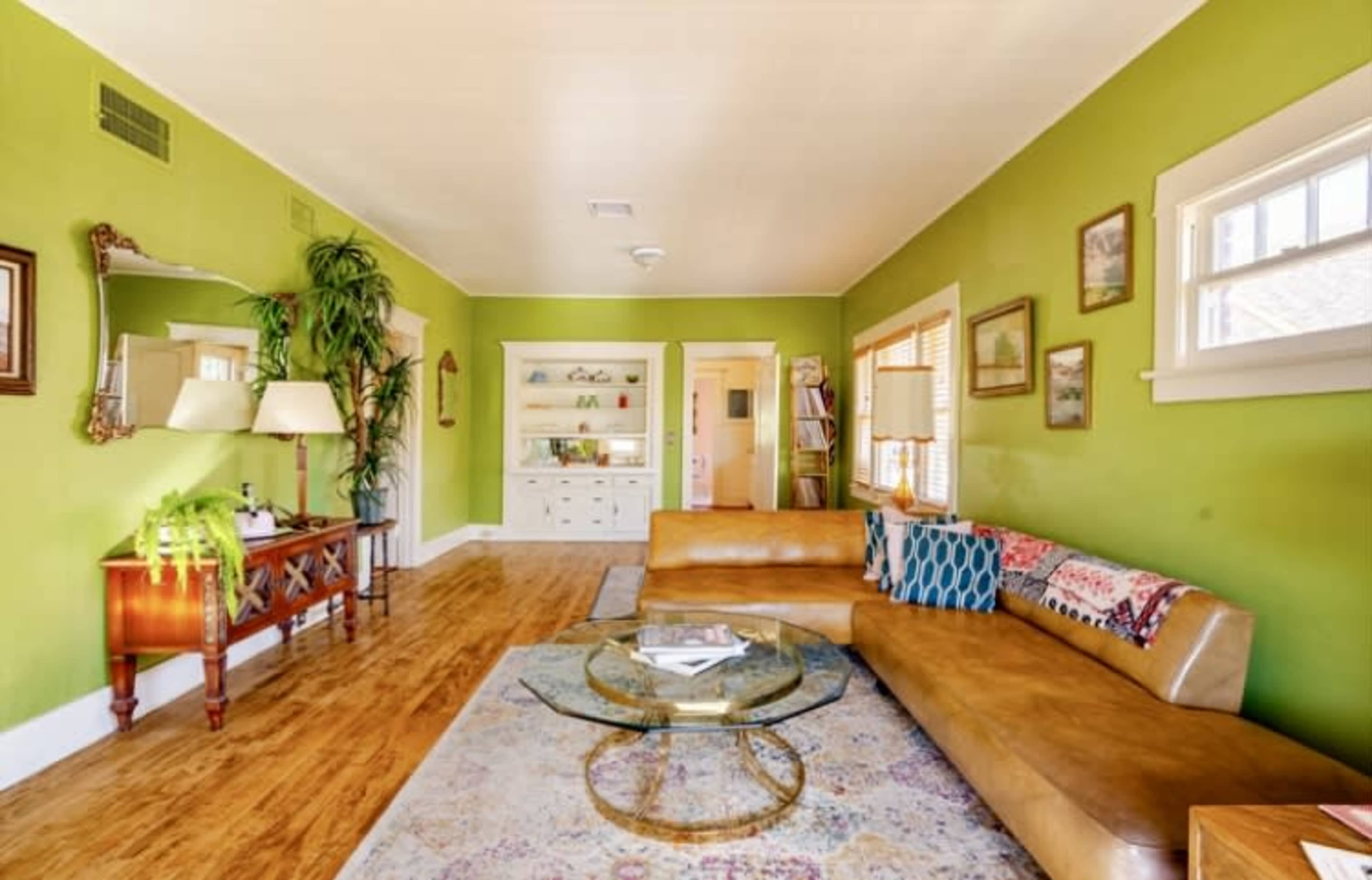 A bright green living room features a brown leather sectional sofa, a round glass coffee table, and wooden flooring with a decorative area rug.