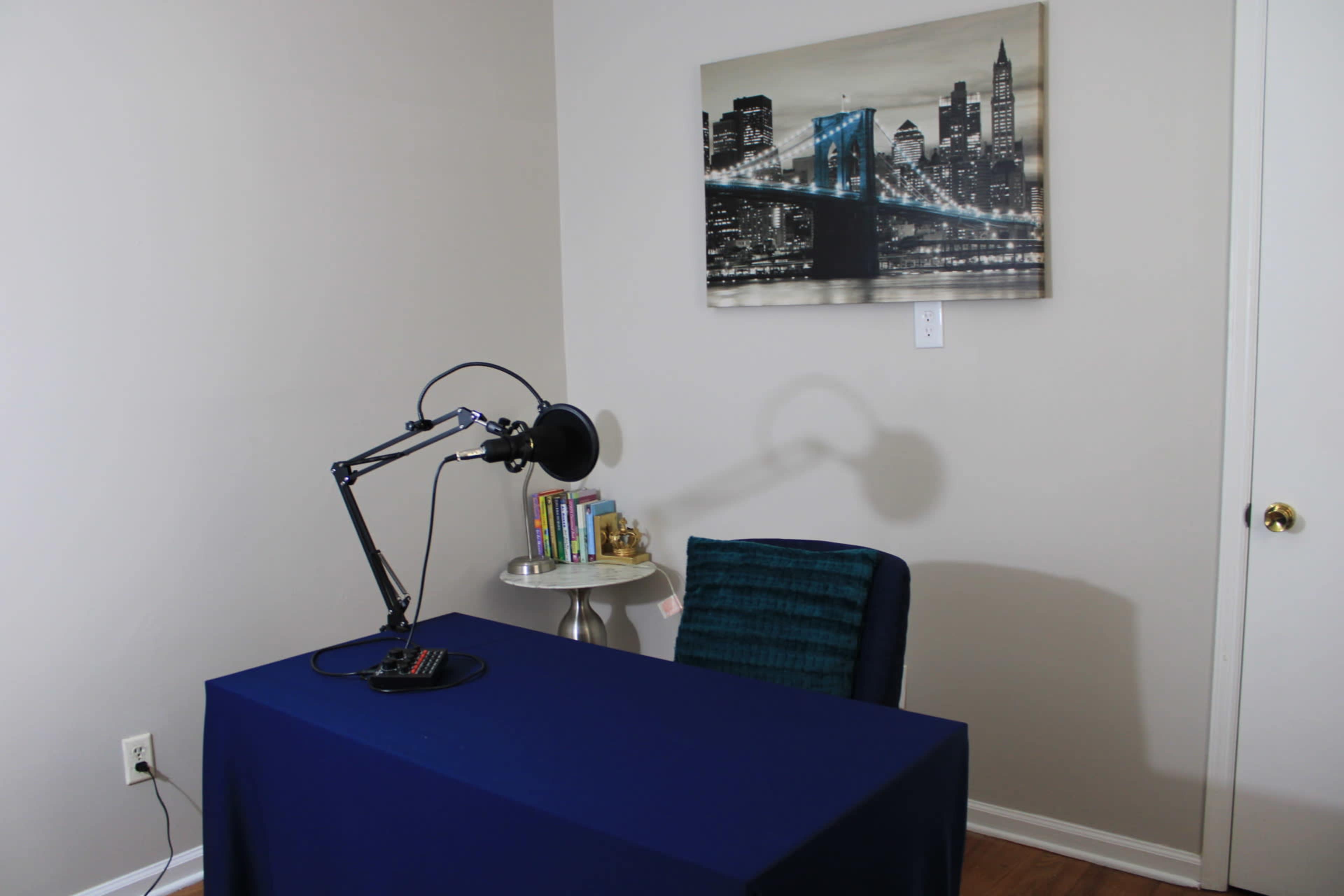 The image shows a simple office space featuring a table covered with a blue cloth, a chair, a lamp on an adjustable arm, and a wall-mounted picture of a city skyline.