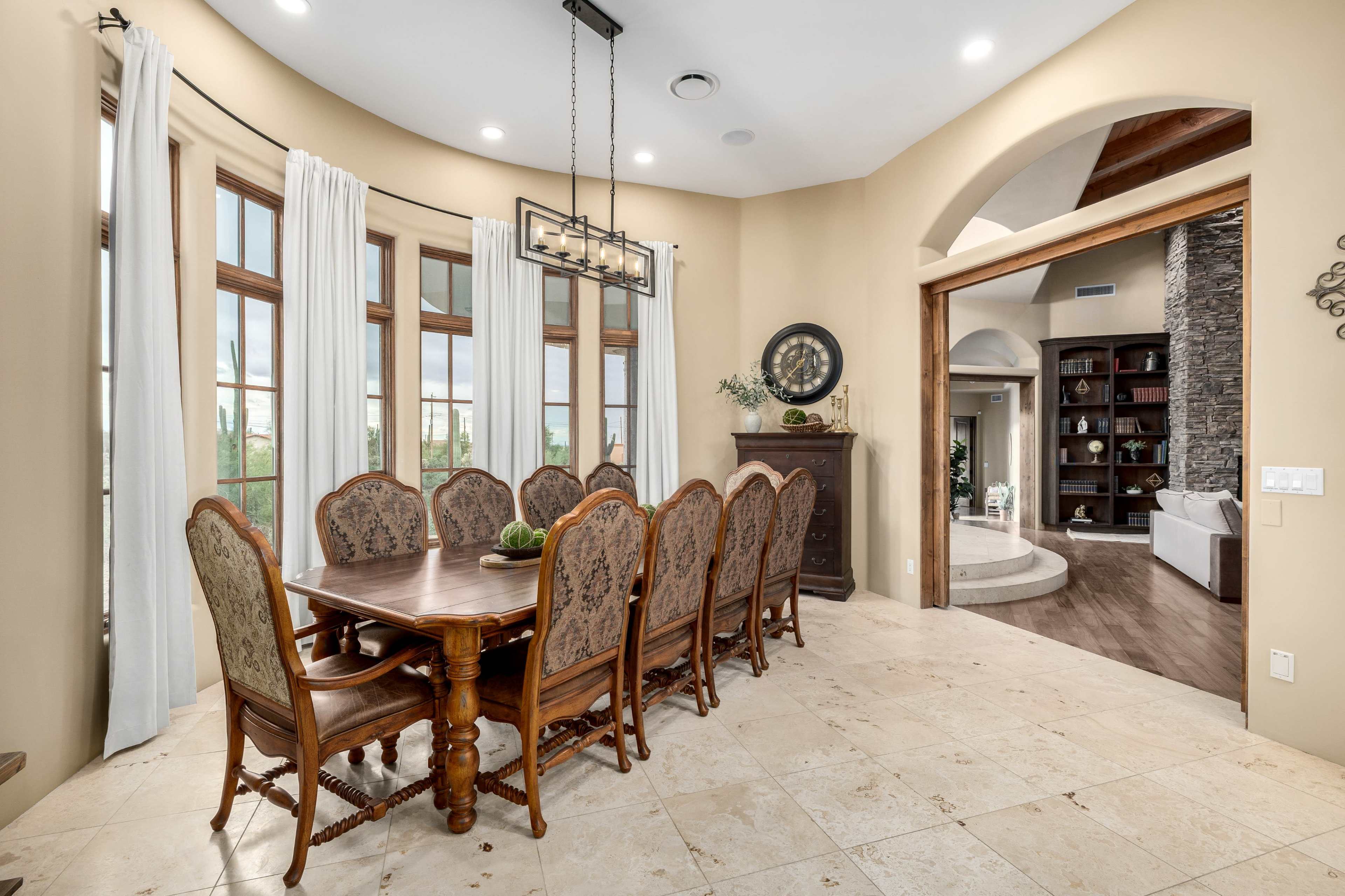 A large dining room features an oval table surrounded by eight upholstered chairs, with tall windows allowing natural light and a view of the outdoors.