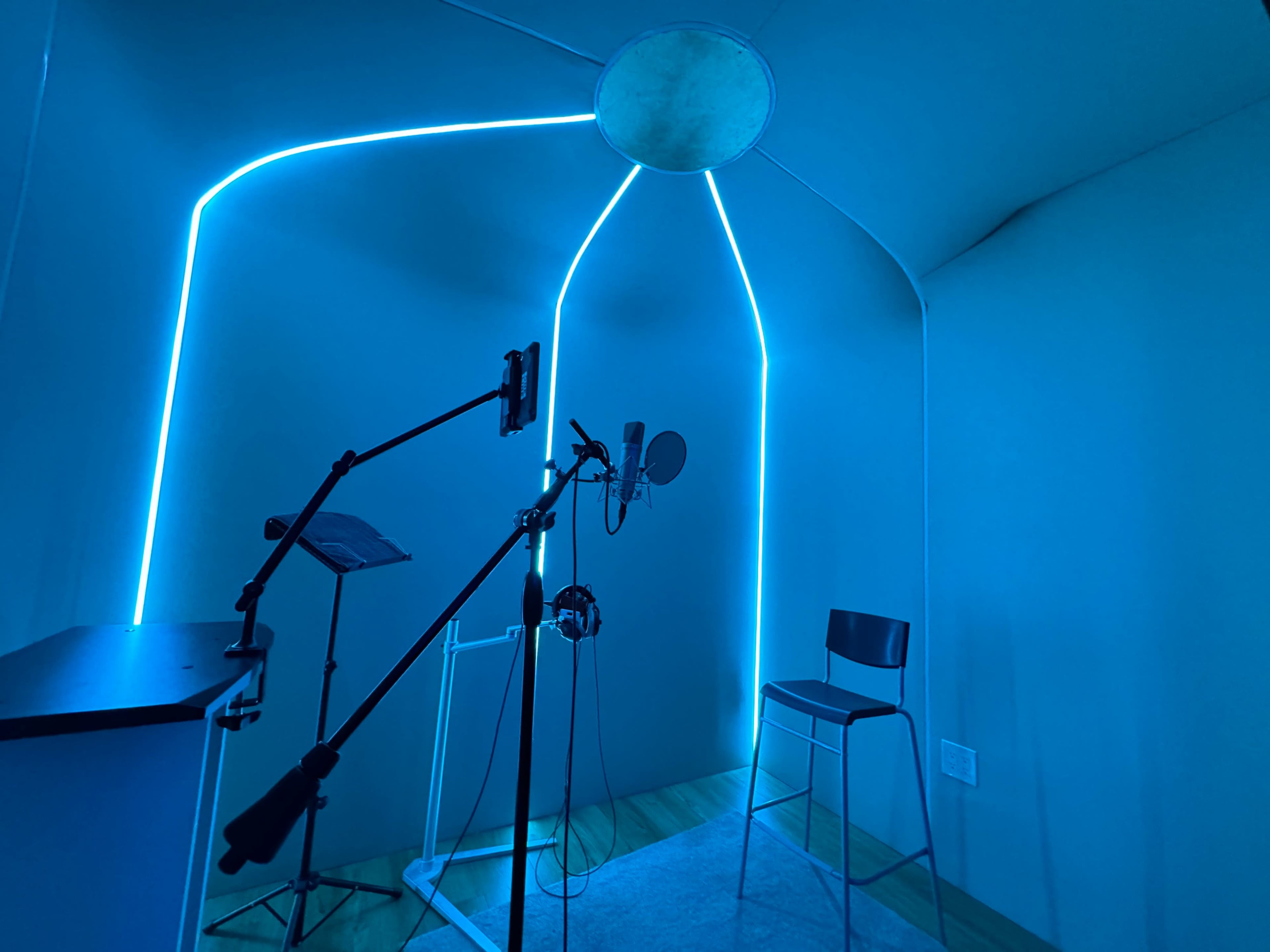 A sound recording booth with blue LED lighting, a microphone on a stand, headphones, and a metal chair.