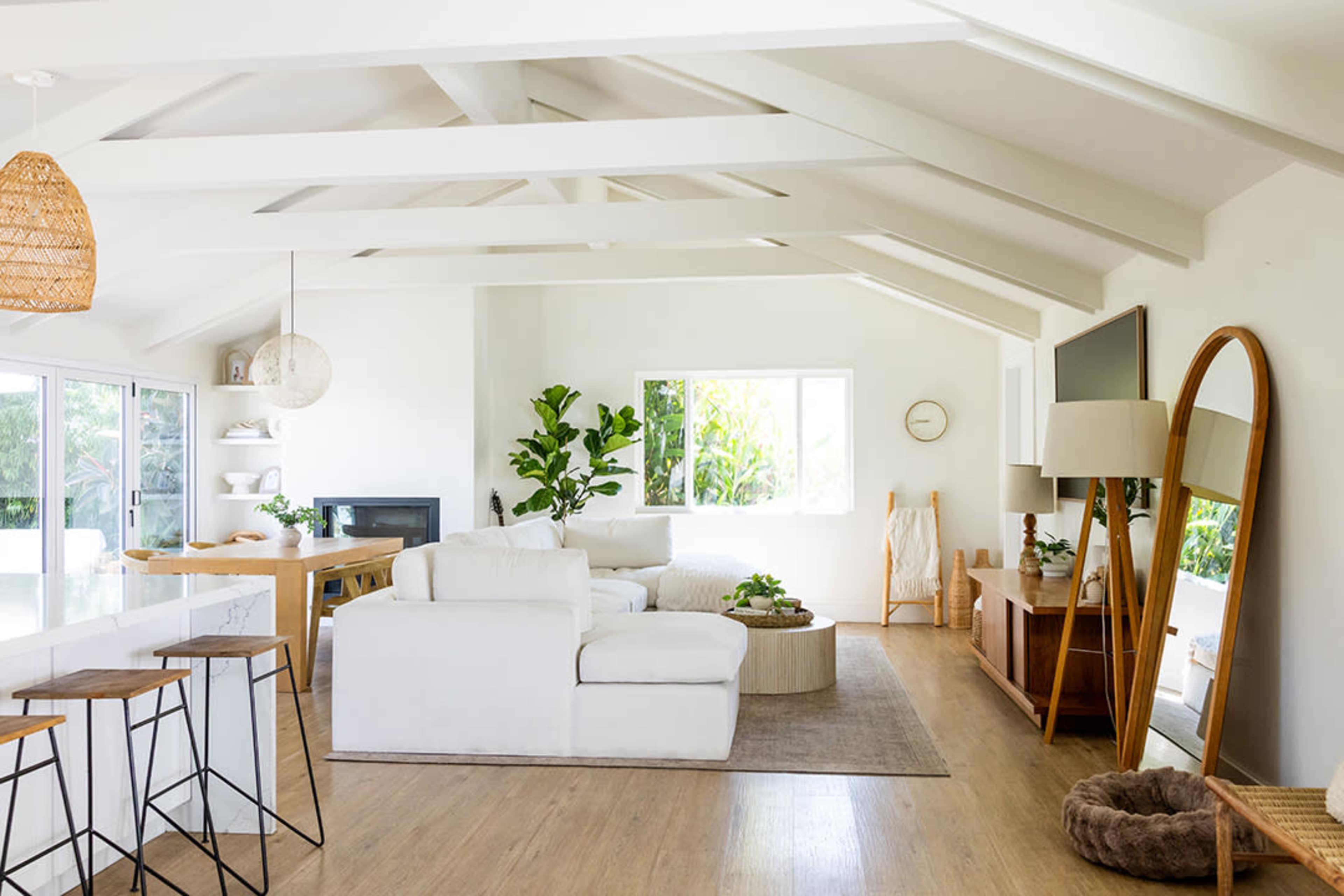 The image shows a bright, modern living room with white walls, a large white sectional sofa, wooden furniture, and greenery visible through the windows.