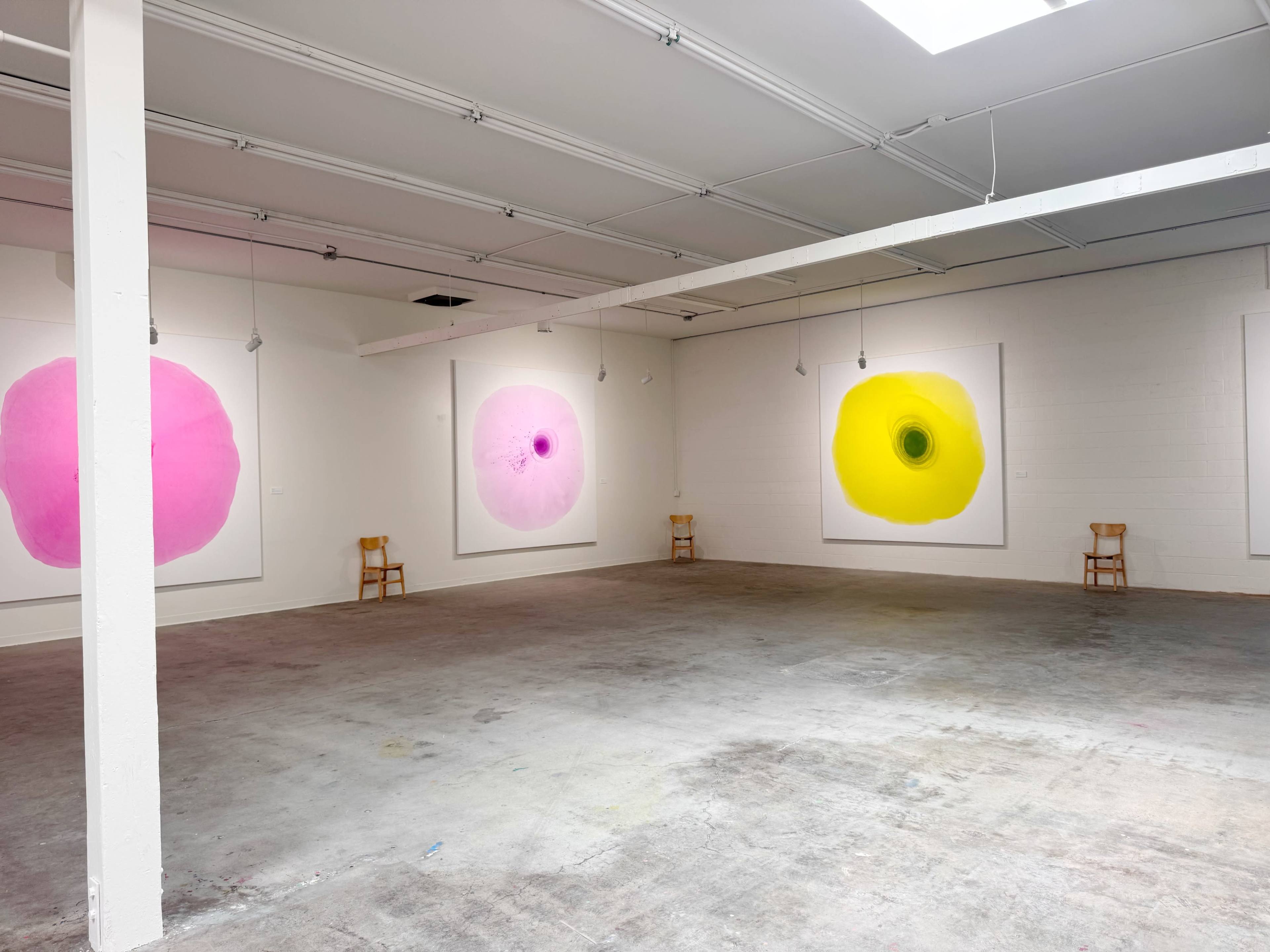 The image shows an art gallery with four large circular paintings in pink, purple, and yellow displayed on the walls, and two wooden chairs placed in the empty space.