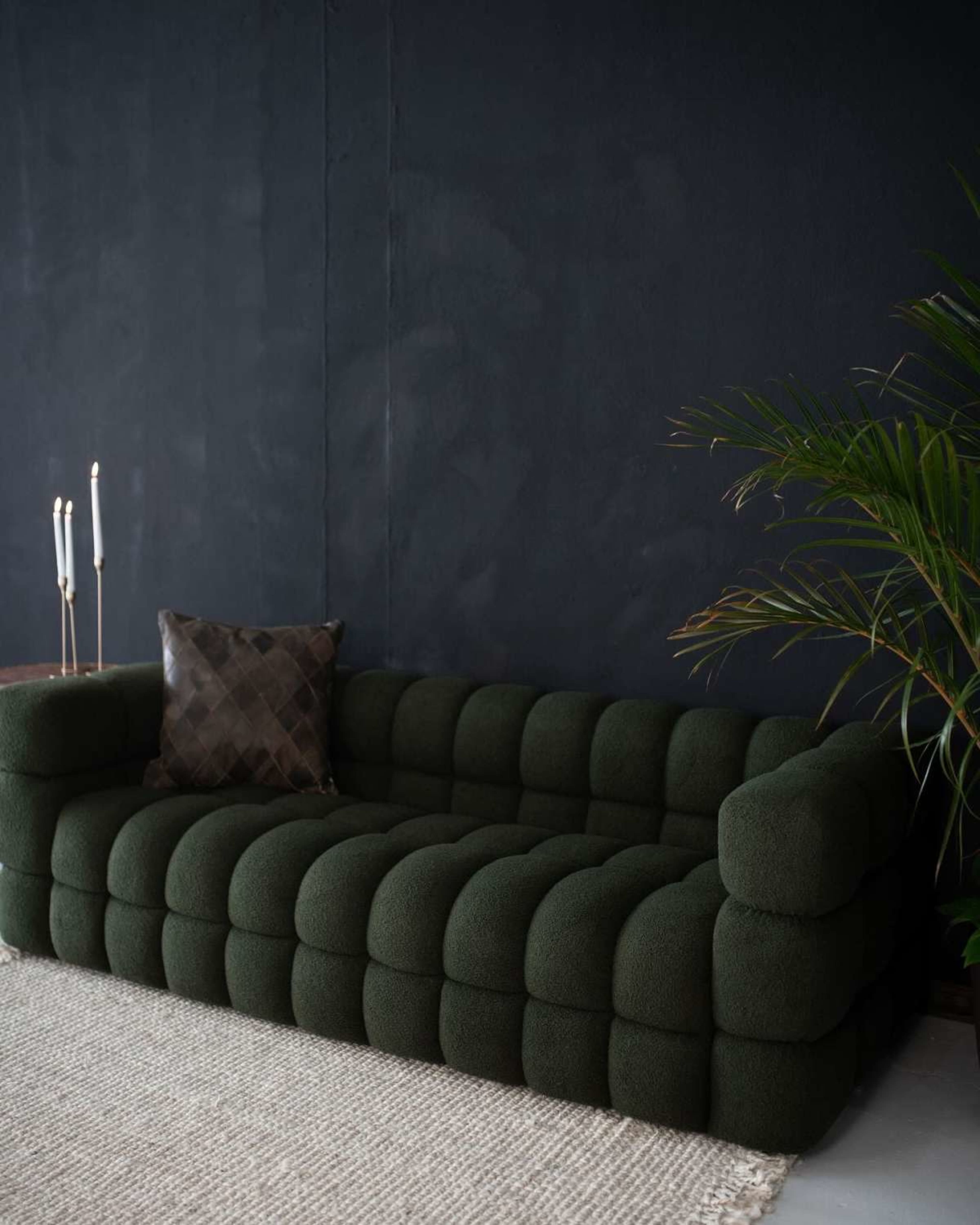A green, upholstered sofa with a rounded design is positioned against a dark wall, accompanied by a decorative pillow and a nearby plant.