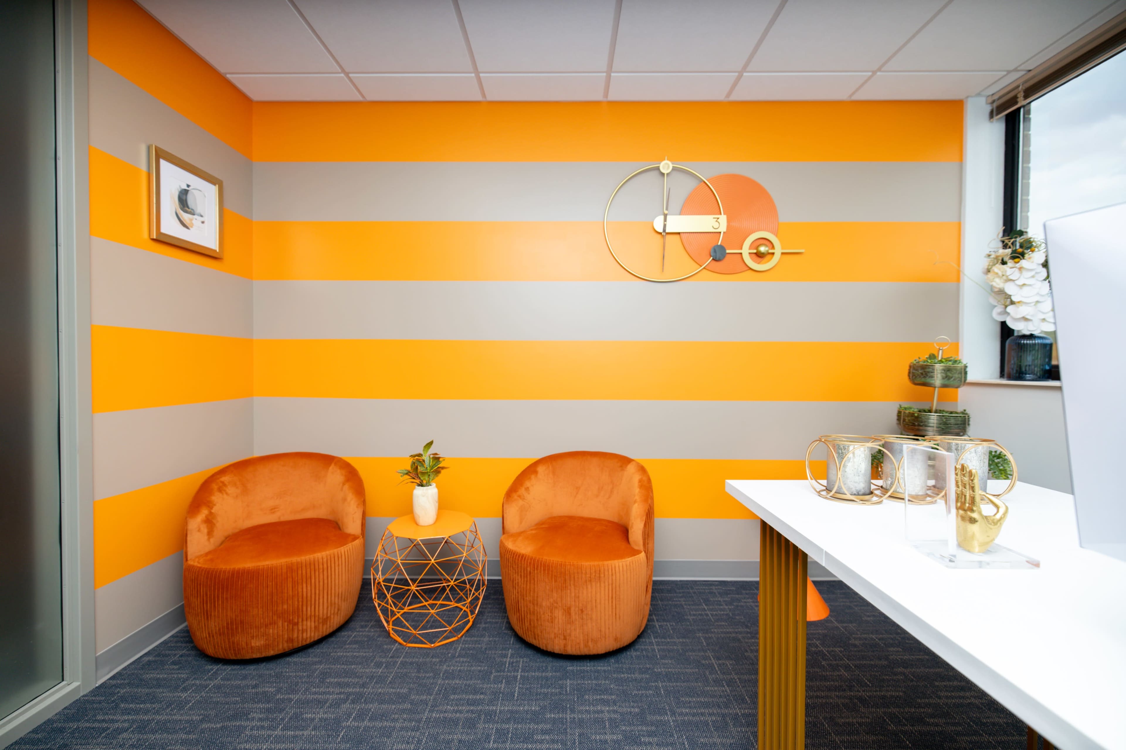A modern office space with orange and gray striped walls, two round orange chairs, a geometric table, and a white desk in the foreground.