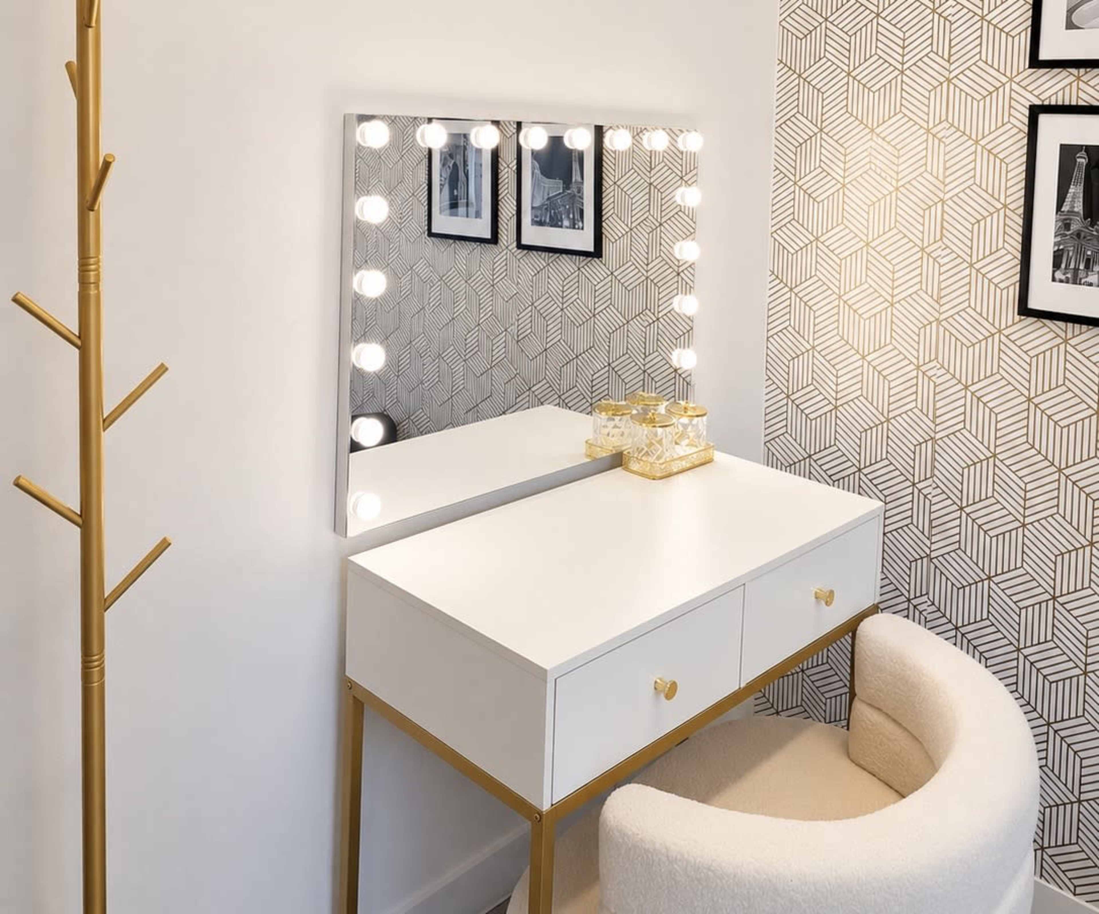 The image shows a modern vanity table with a large illuminated mirror, two drawers, and a circular upholstered chair set against a patterned wall.