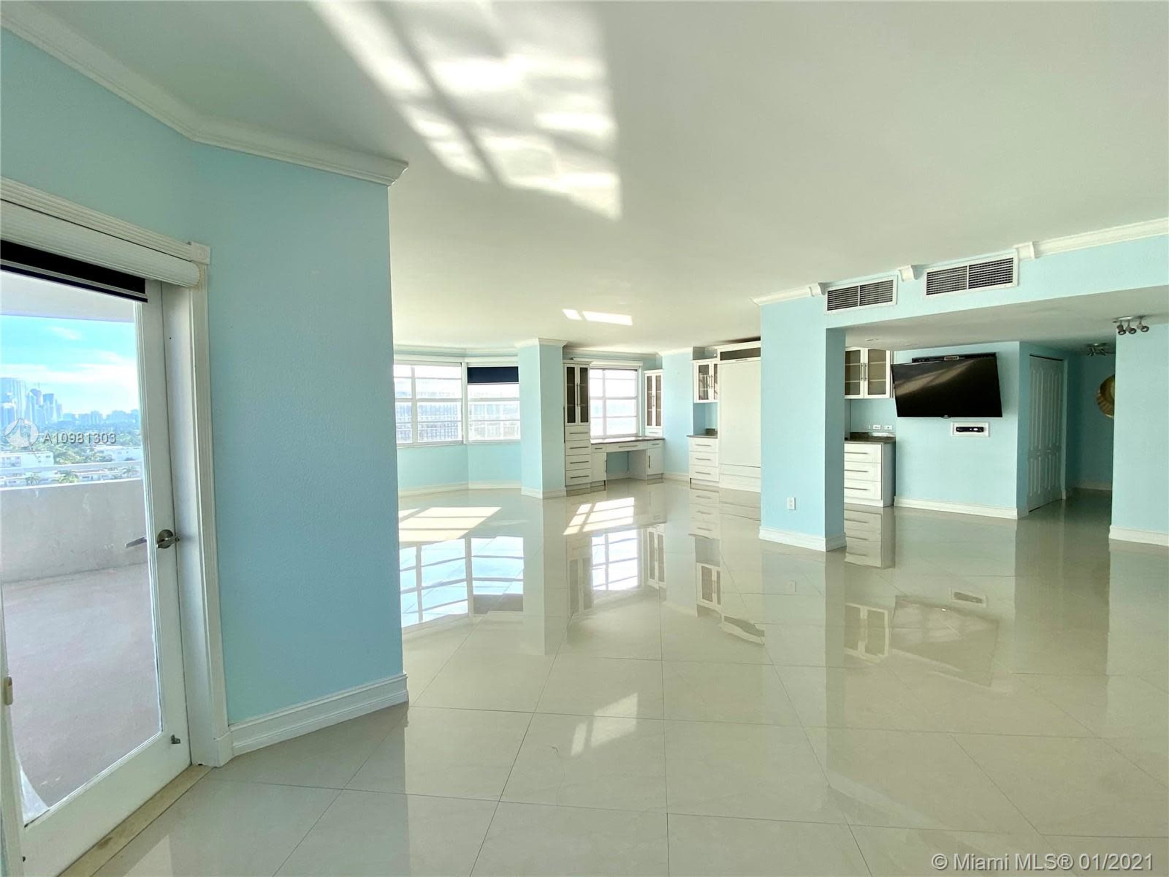 A spacious, light-filled living area with pale blue walls and glossy tile flooring, featuring large windows and built-in cabinetry.