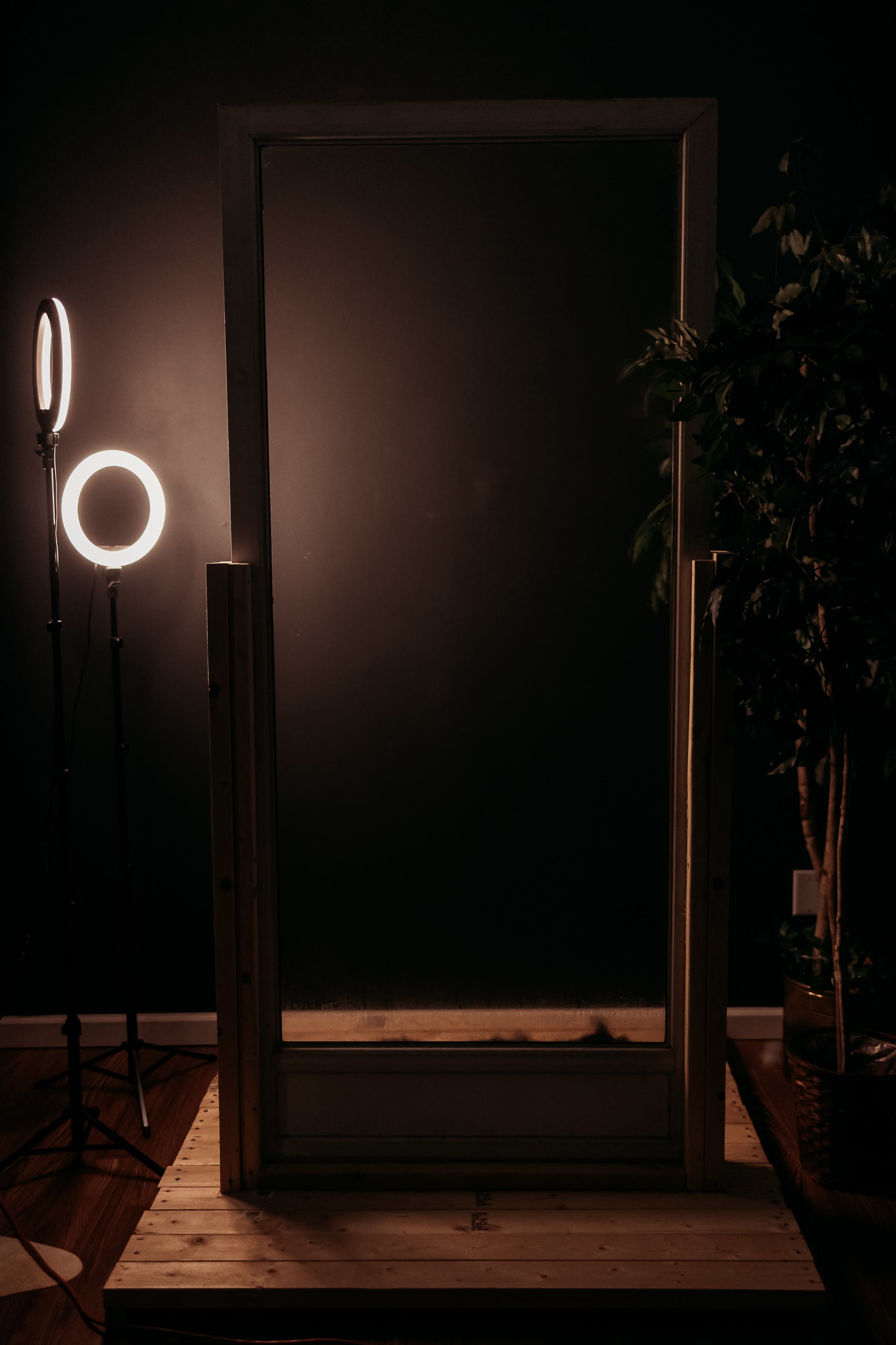A wooden frame with a large mirror stands on a wooden base, illuminated by a round light source and accompanied by a plant in the background.