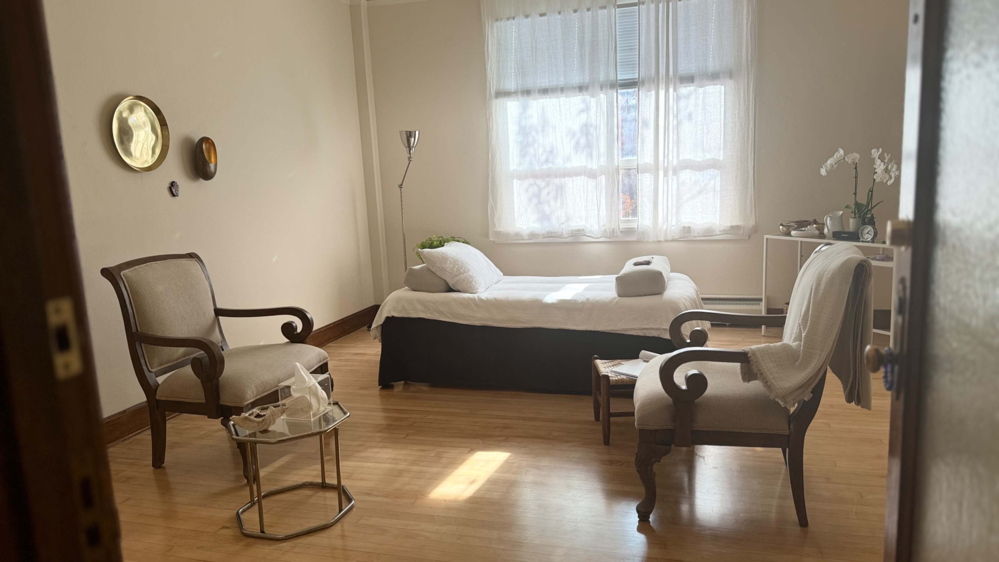 The image shows a minimalist room with a bed, two chairs, a small table, and a lamp, featuring large windows that let in natural light.