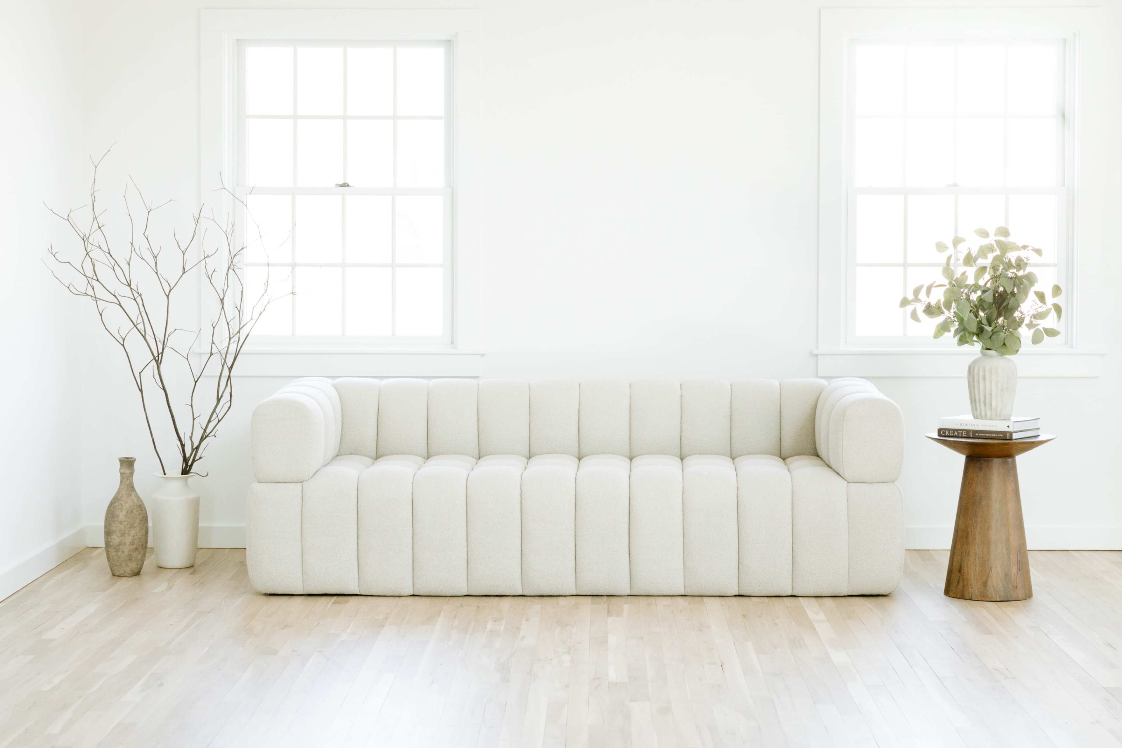 A light-colored, tufted sofa is positioned against a white wall in a minimalist room with large windows, accompanied by a decorative vase and a small table with a plant.