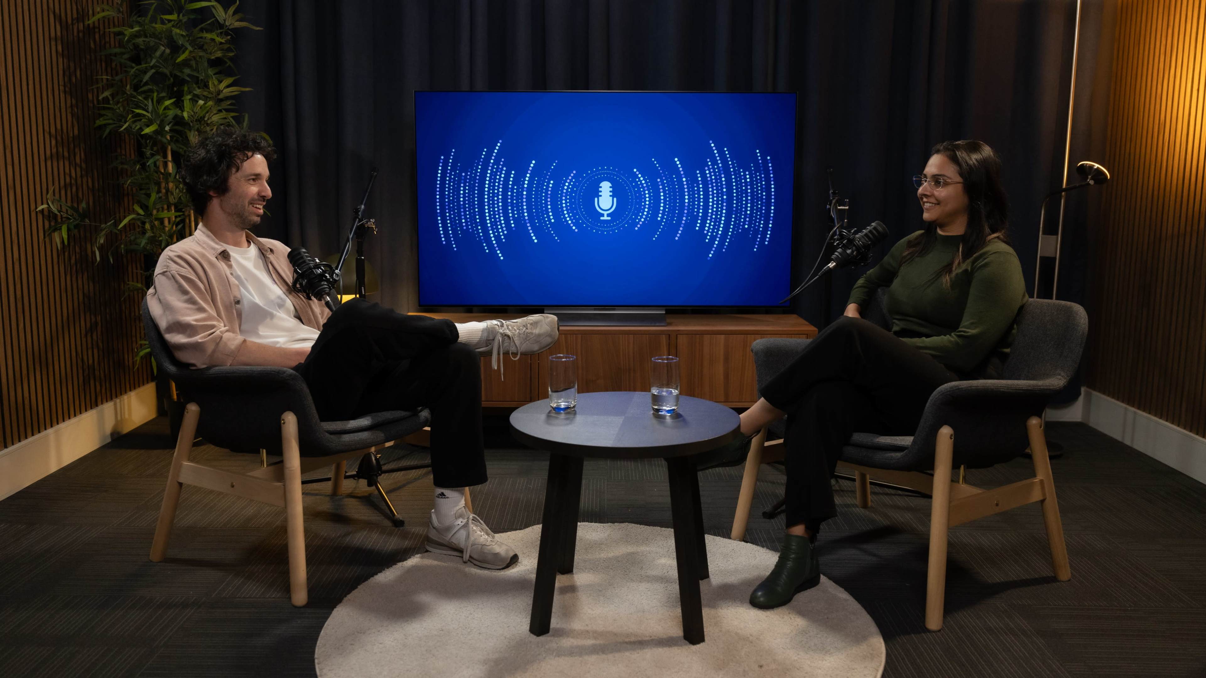 A man and a woman are seated at a small table in a modern studio, with microphones in front of them and a blue screen displaying sound waves in the background.