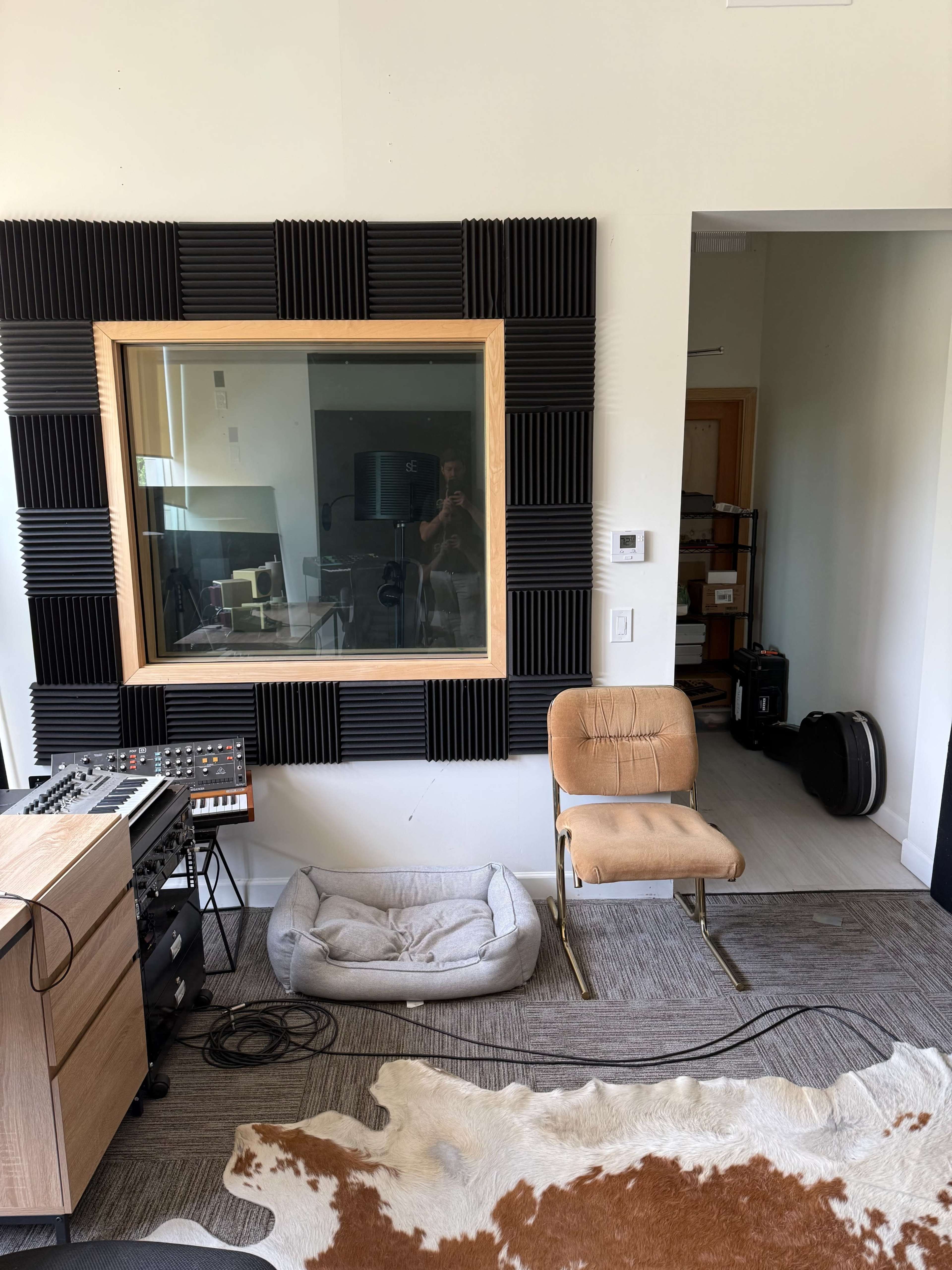 The image shows a soundproof studio space featuring a large glass window, a wooden console with electronic equipment, a chair, and a pet bed on a carpeted floor.