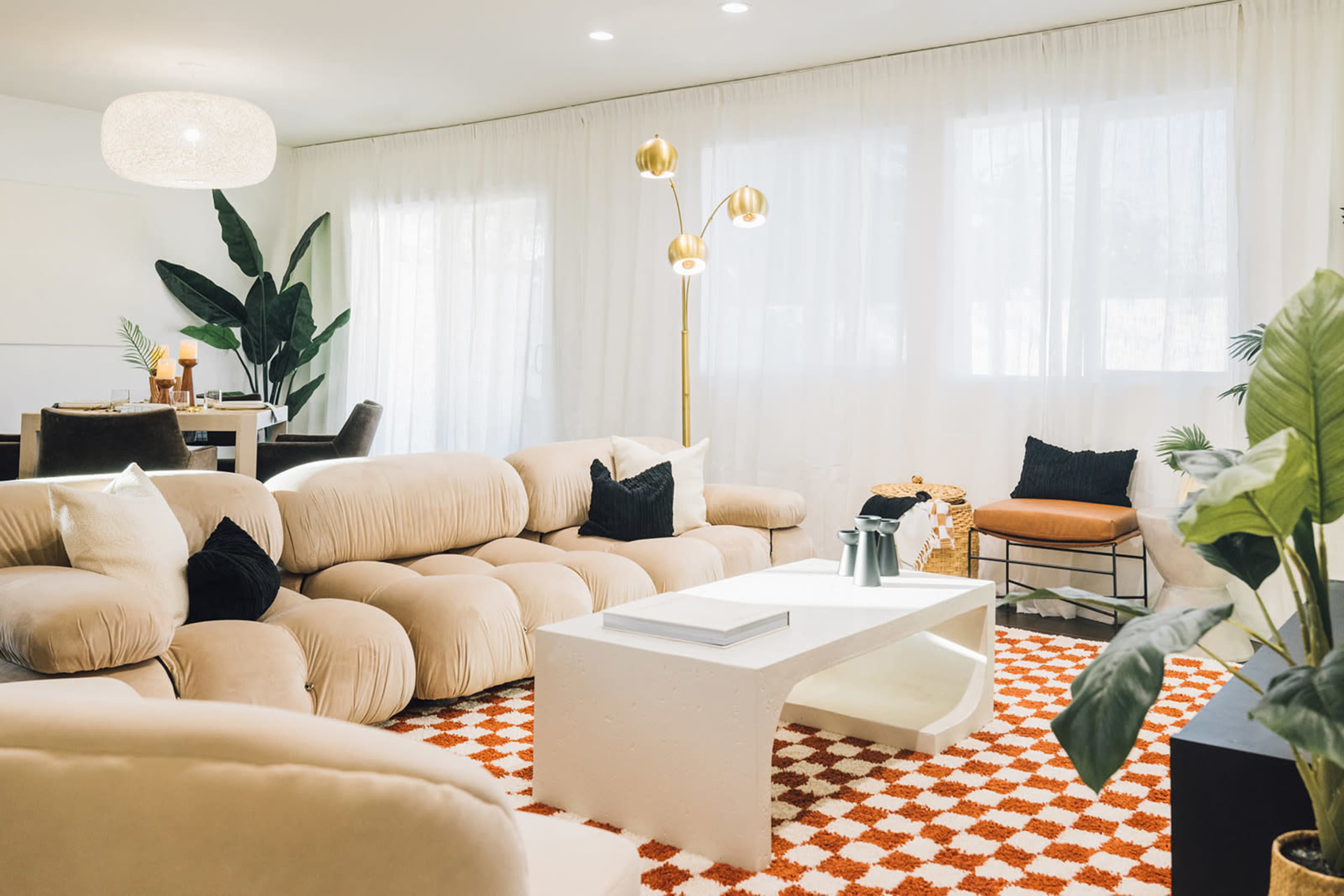 A modern living room with a beige, rounded sofa, a white coffee table, and large windows covered by sheer curtains, complemented by indoor plants and a stylish floor lamp.