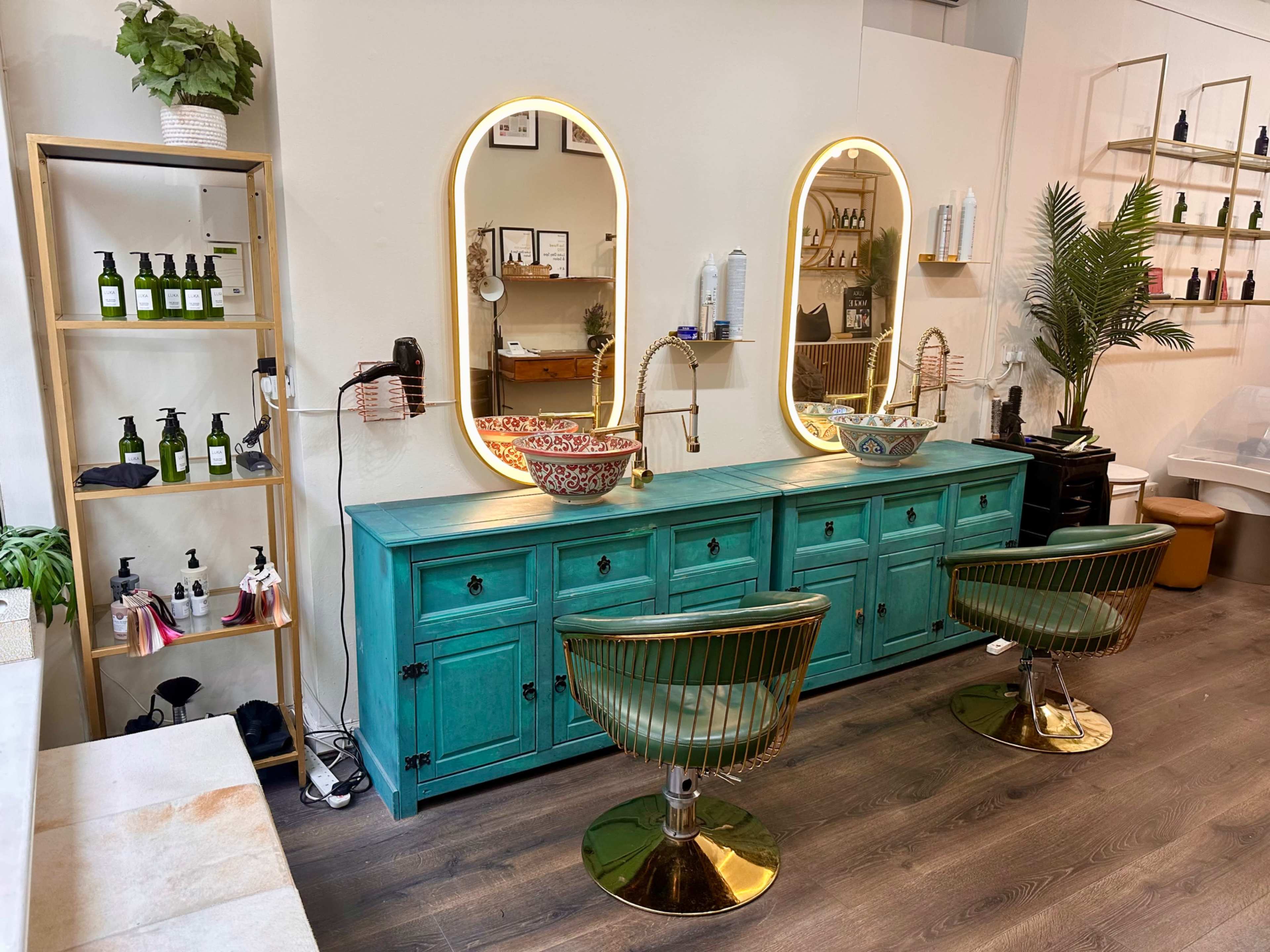 The image shows a brightly colored turquoise salon vanity with two round mirrors and modern gold-accented chairs, surrounded by various hair care products on display.
