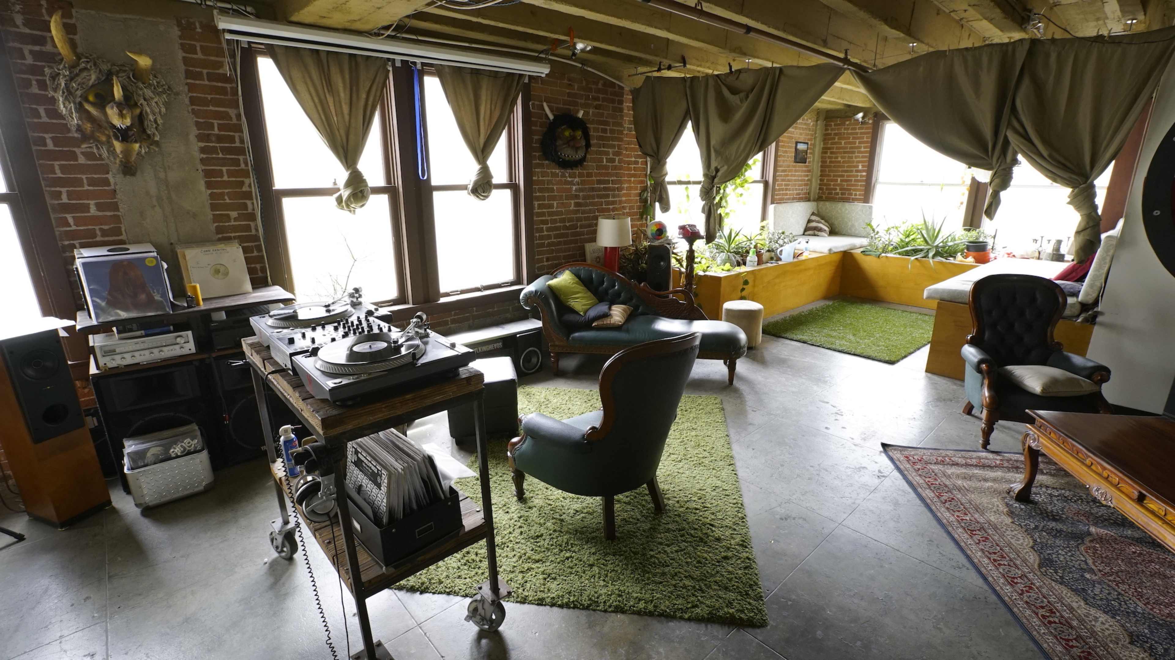 The spacious room features large windows with draped curtains, a vinyl record player on a cart, and a mix of seating arrangements along with plants and a soft green rug.