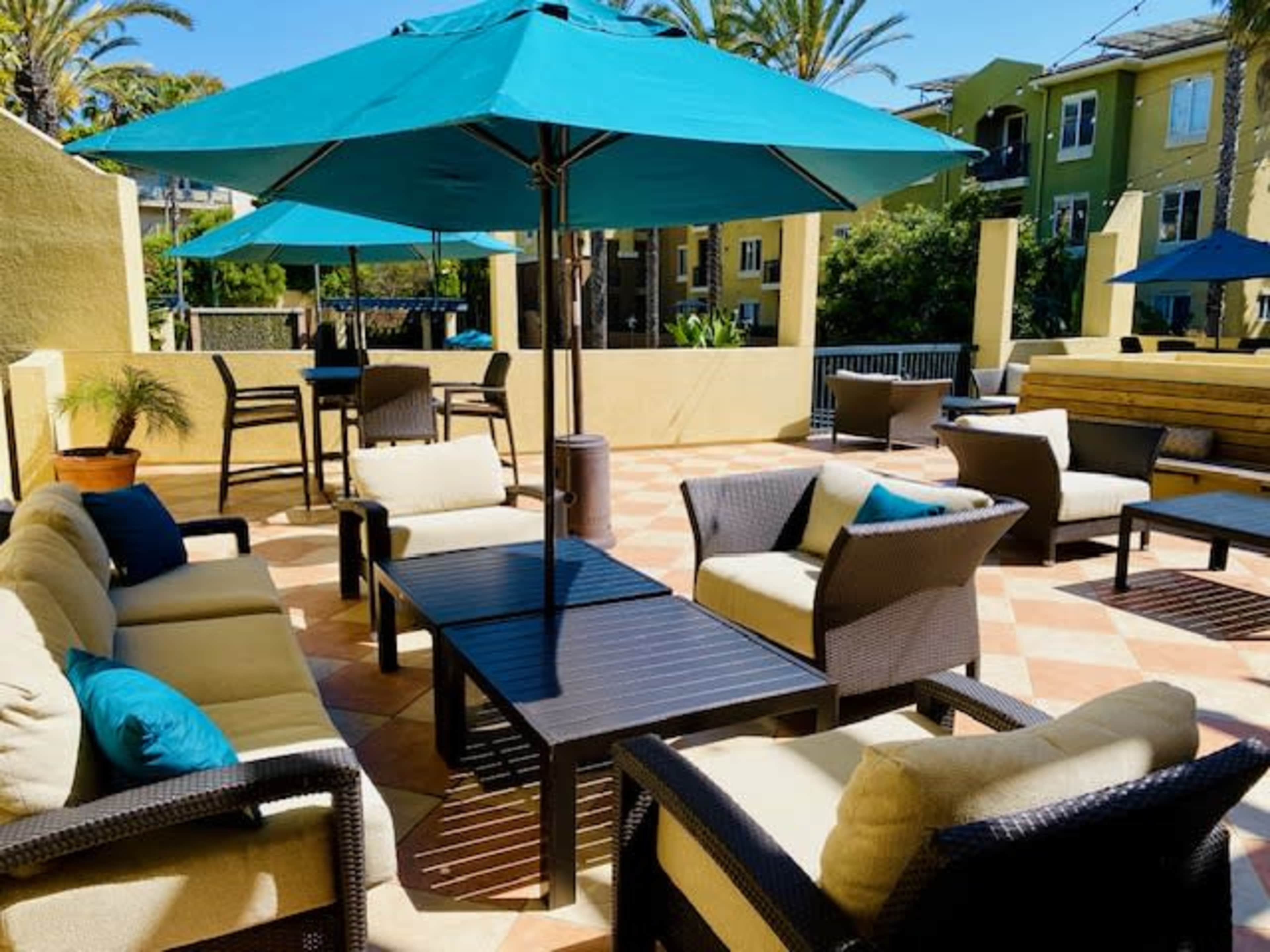 An outdoor lounge area with turquoise umbrellas, wicker seating, and a tiled floor surrounded by palm trees and buildings.