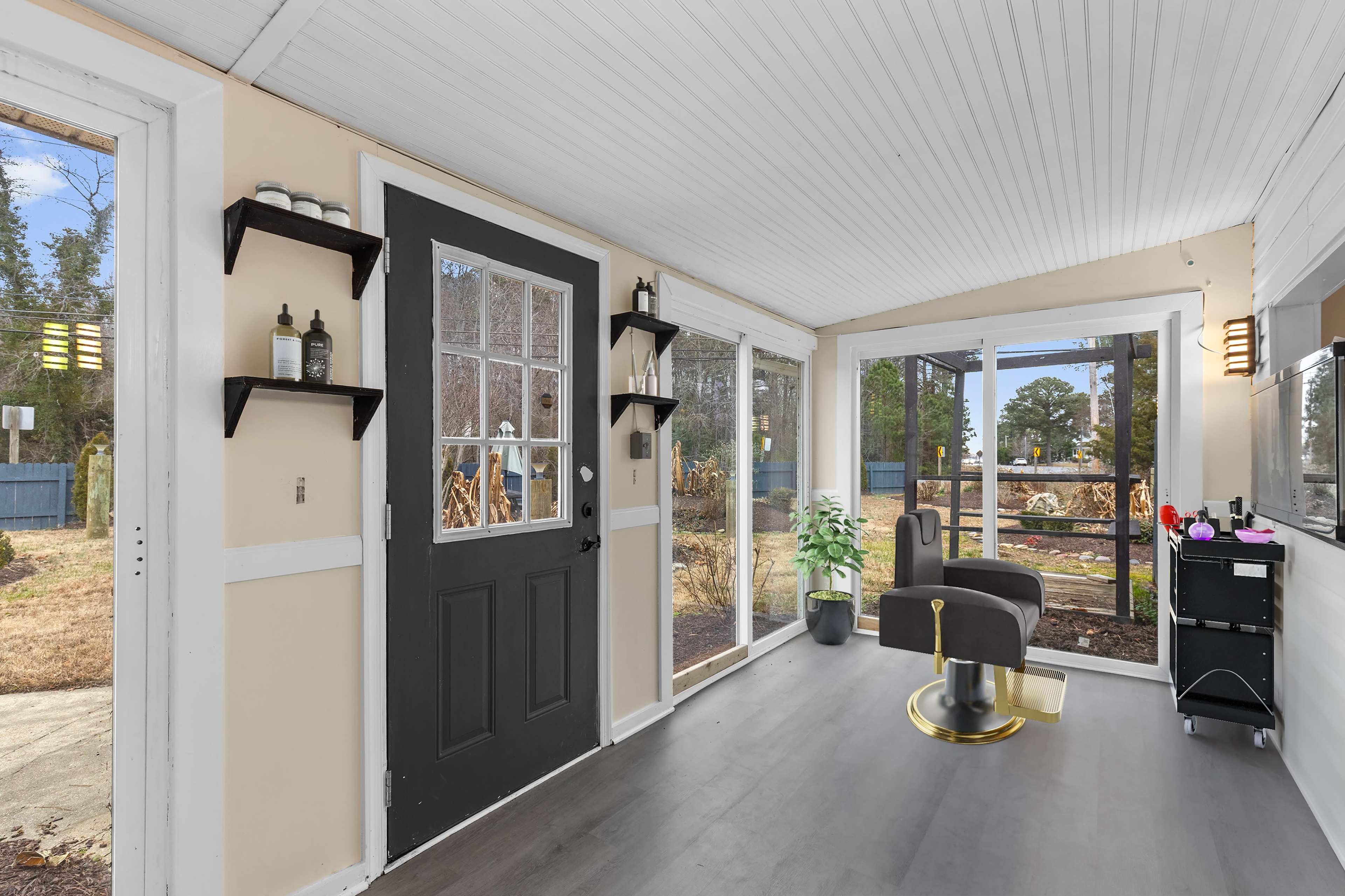 The image shows a sunroom with a black door, large windows, a salon chair, and shelves with bottles on the wall.