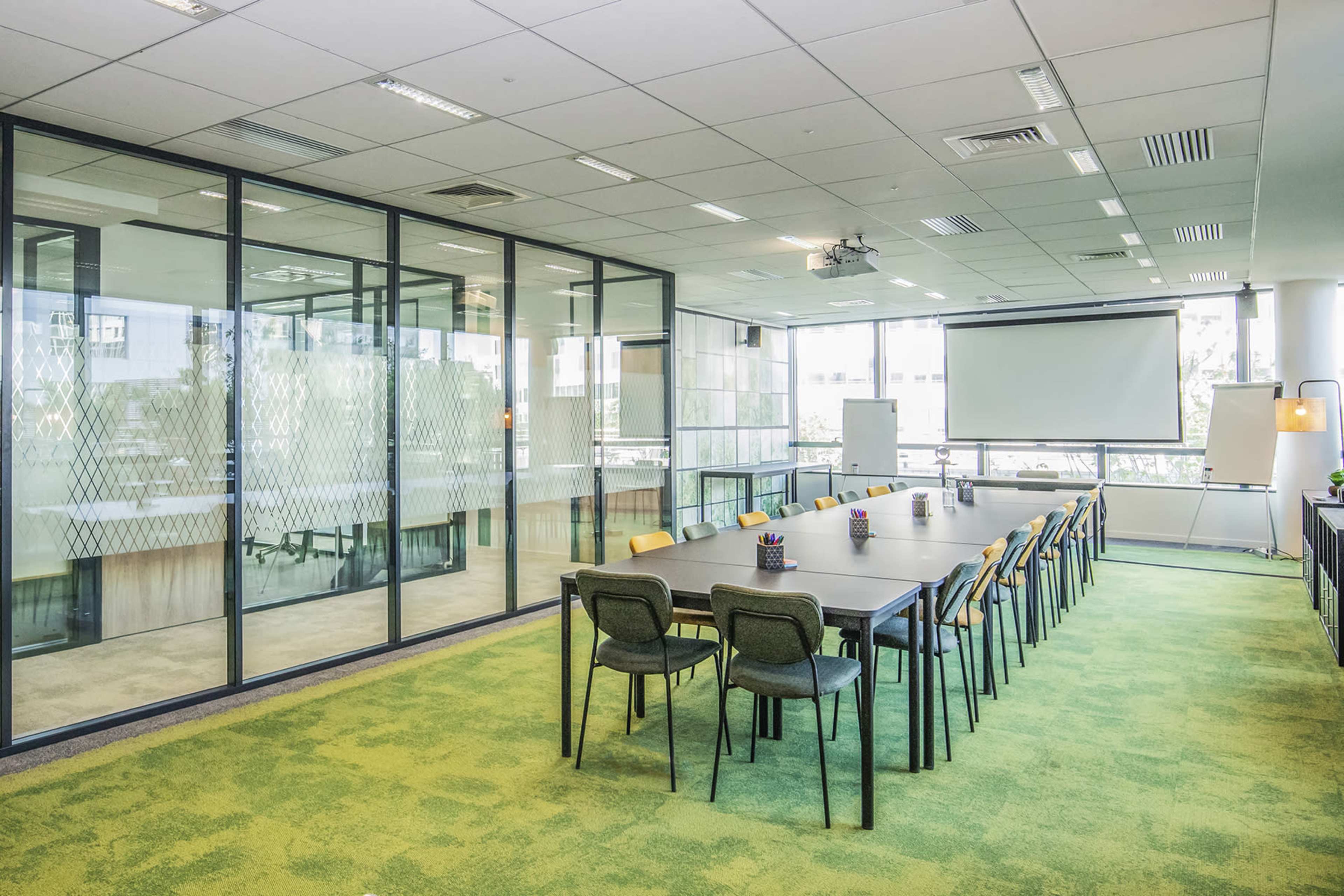 A modern conference room with a long table surrounded by chairs, large windows, and a projector screen at one end.
