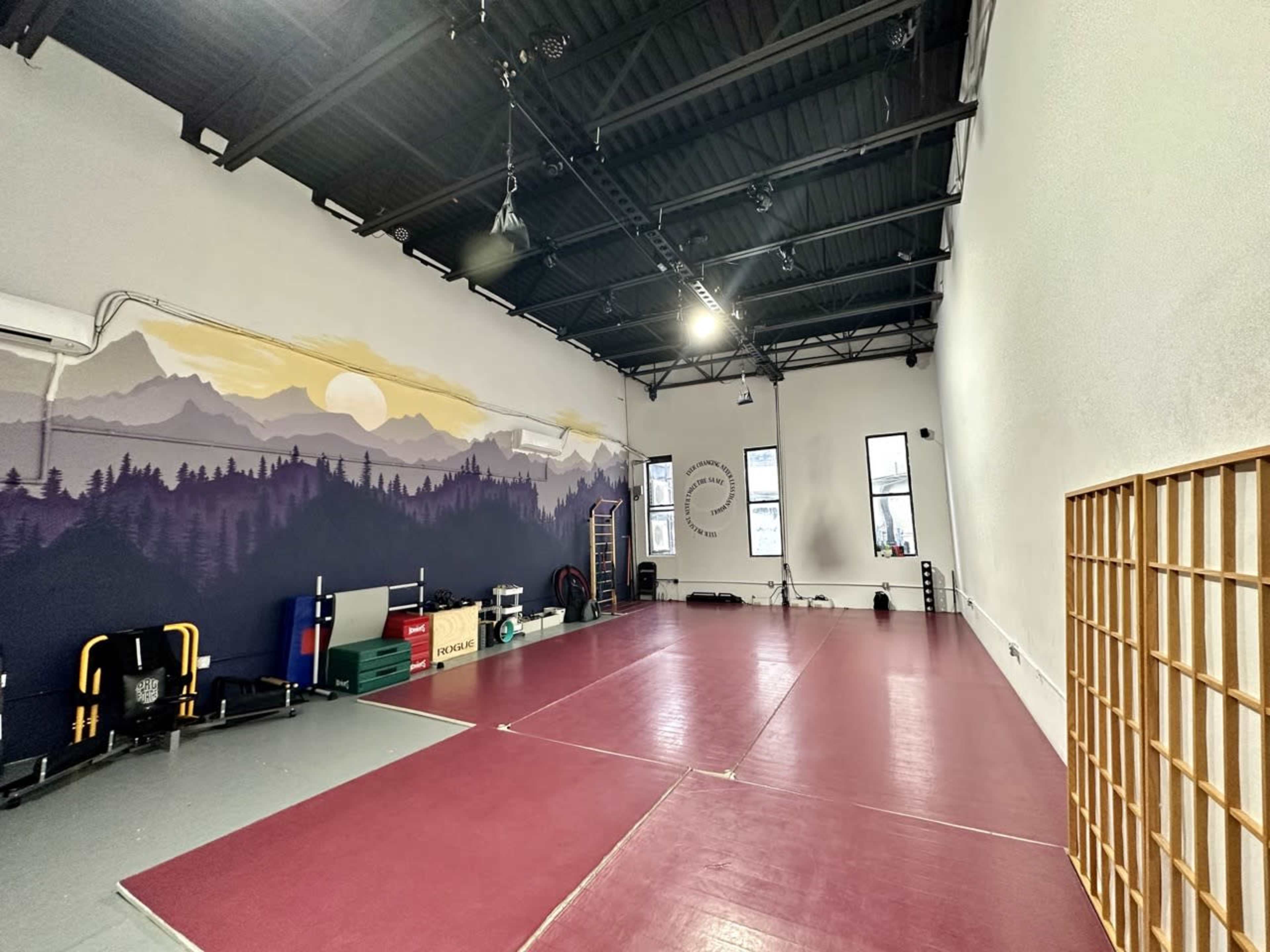 A spacious indoor martial arts studio with a red rubber mat, large windows, and a mural of mountains on one wall.