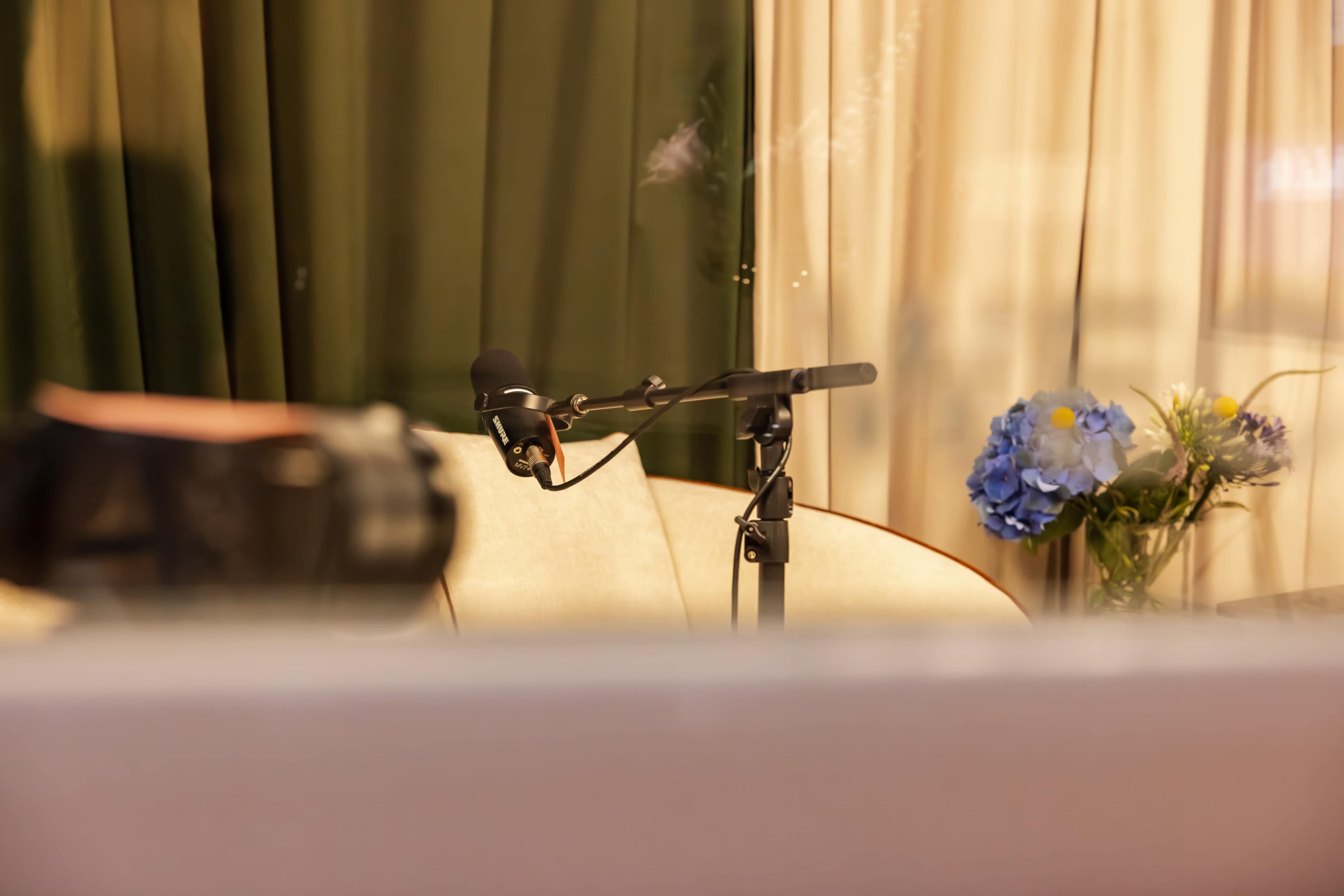 A microphone stands on a black arm in front of a floral arrangement and partially visible furniture, with soft drapes in the background.