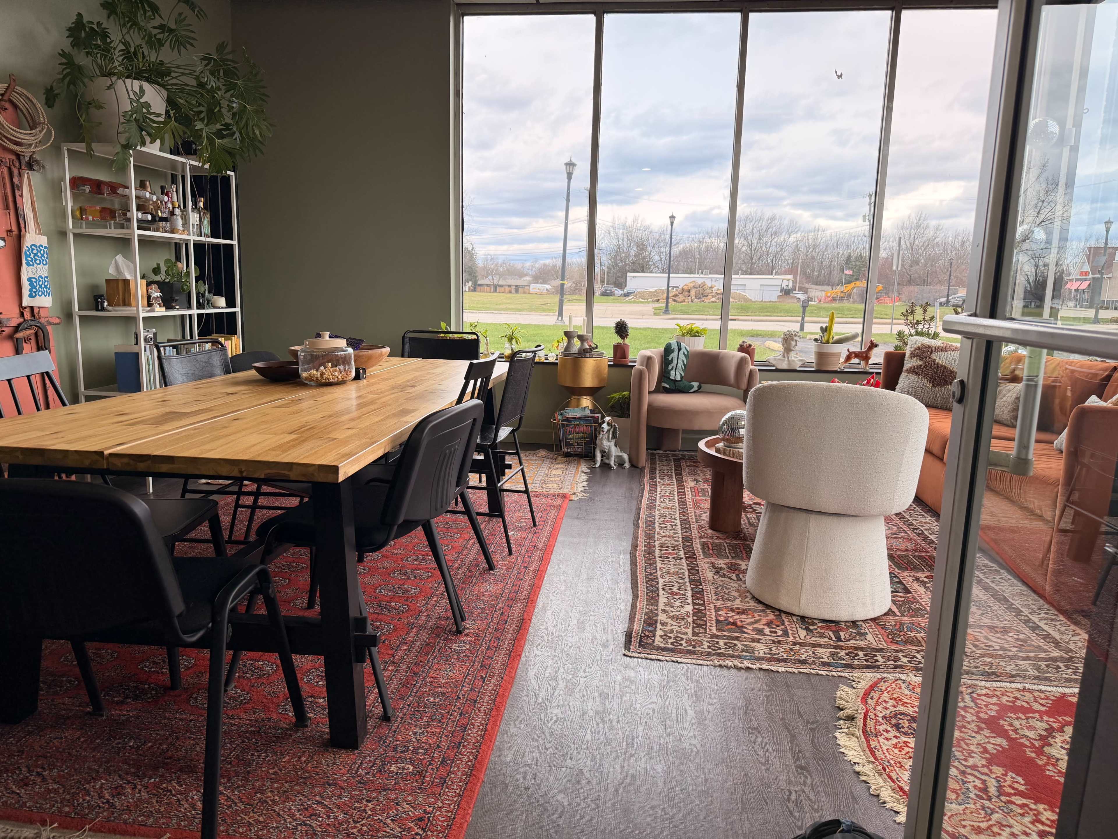 The image shows a well-lit indoor space with a wooden dining table, black chairs, and a seating area featuring a sofa and a white chair, all surrounded by large windows overlooking a grassy area.