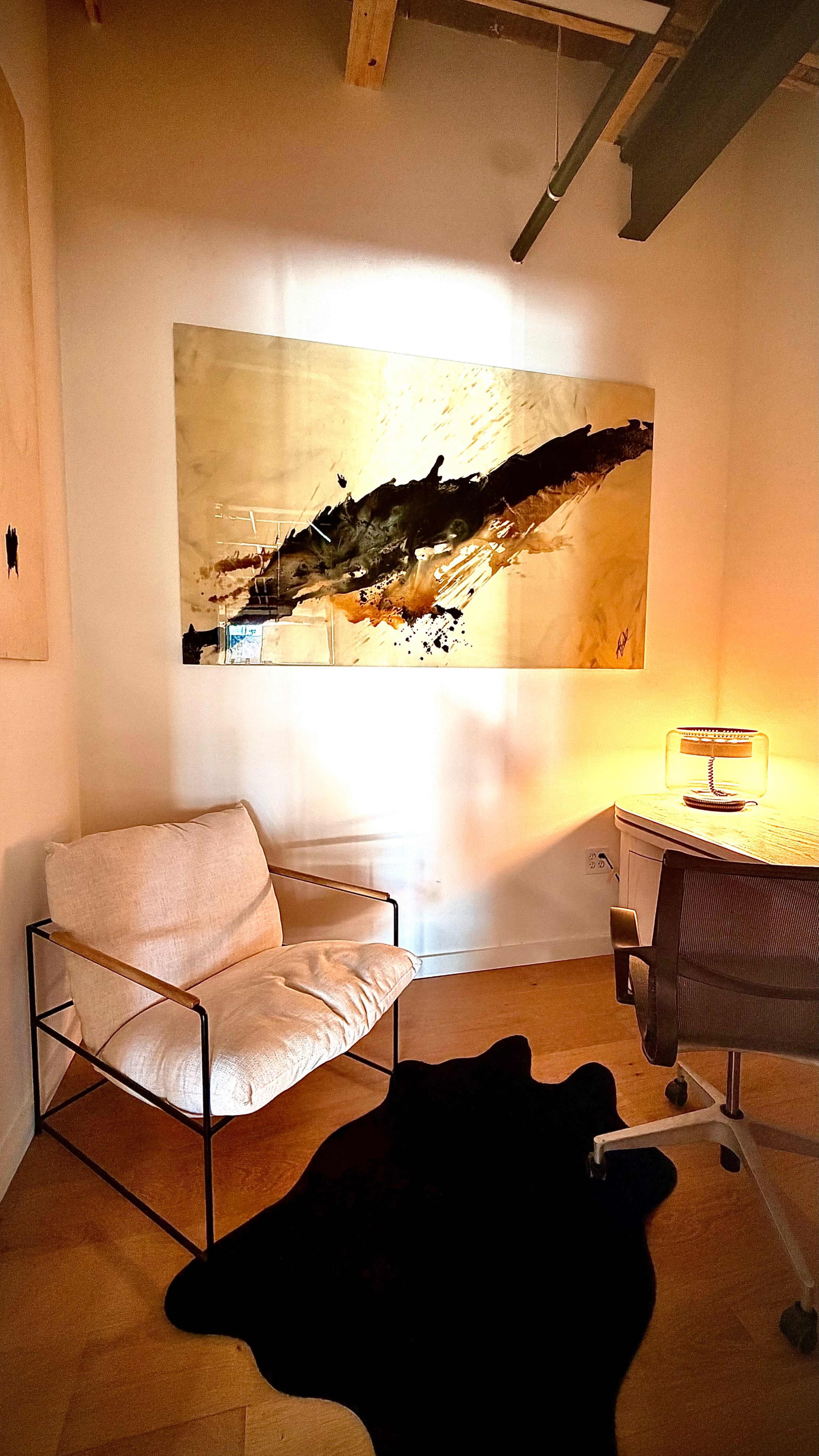 The image shows a modern office space featuring a light-colored armchair, a black rug, a desk with a lamp, and an abstract painting on the wall.