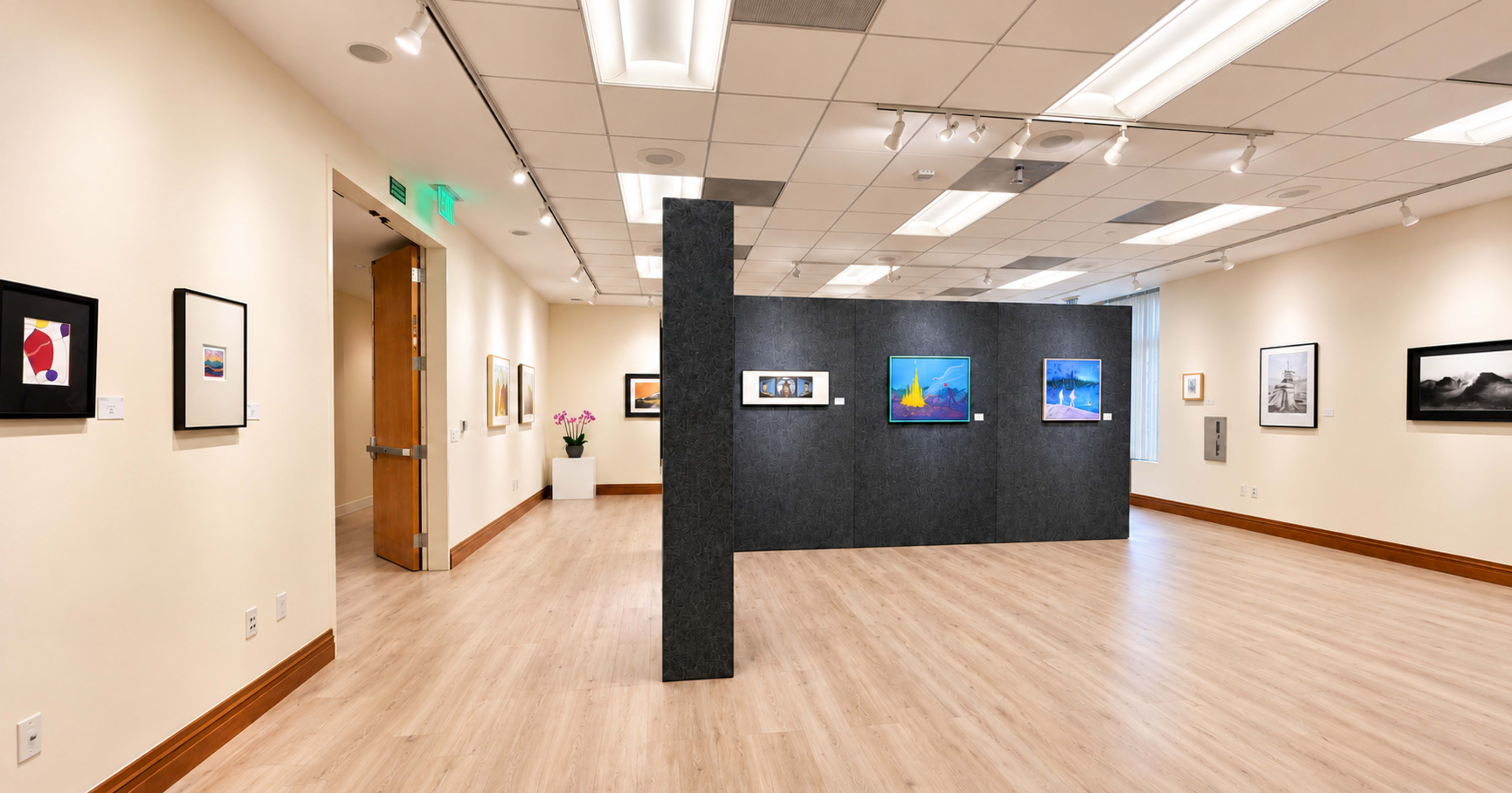 The image depicts a bright art gallery with framed artworks displayed on the walls and a partition separating the space.