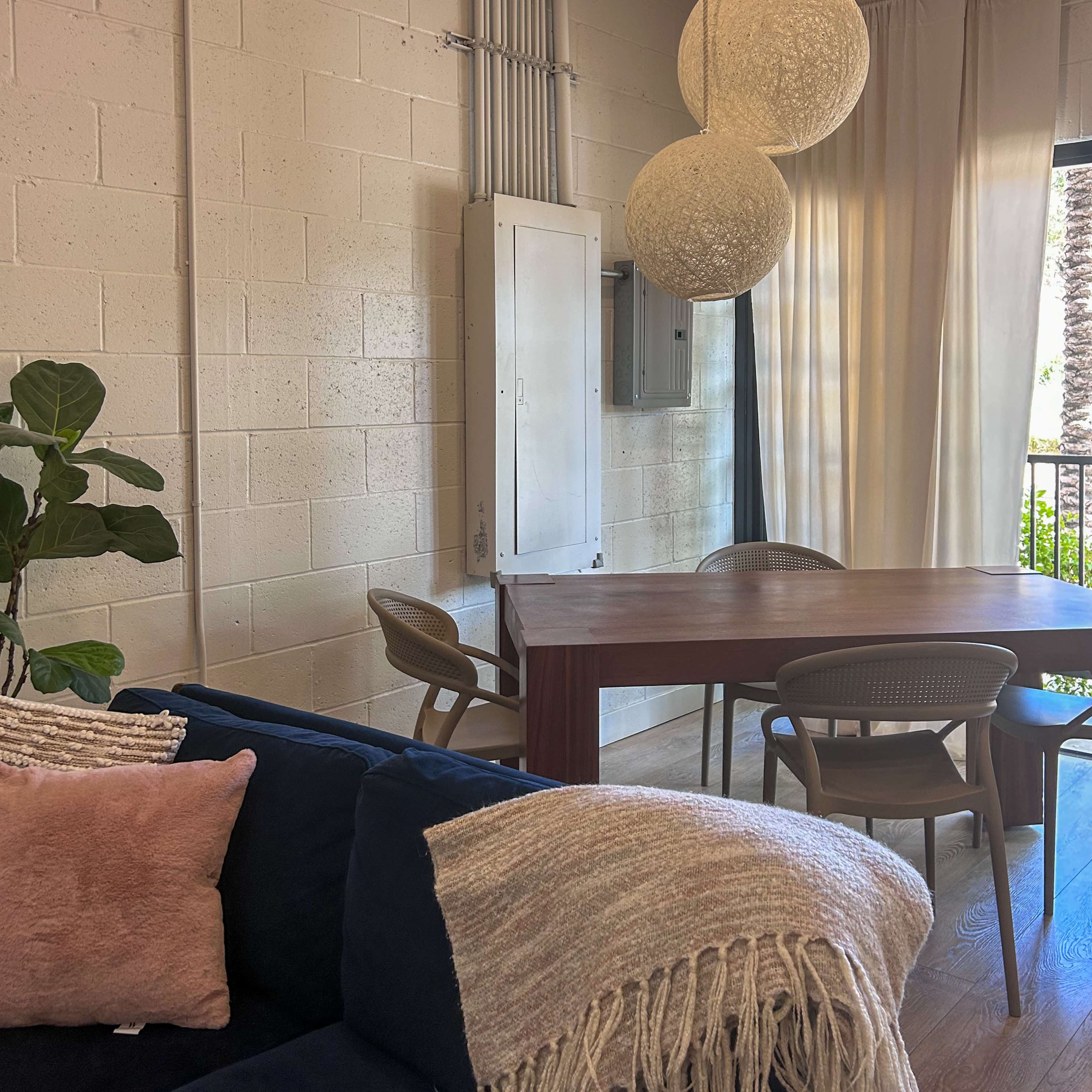 The image shows a modern living space with a dining table, four chairs, a large light fixture, and a sofa with decorative pillows and a blanket.