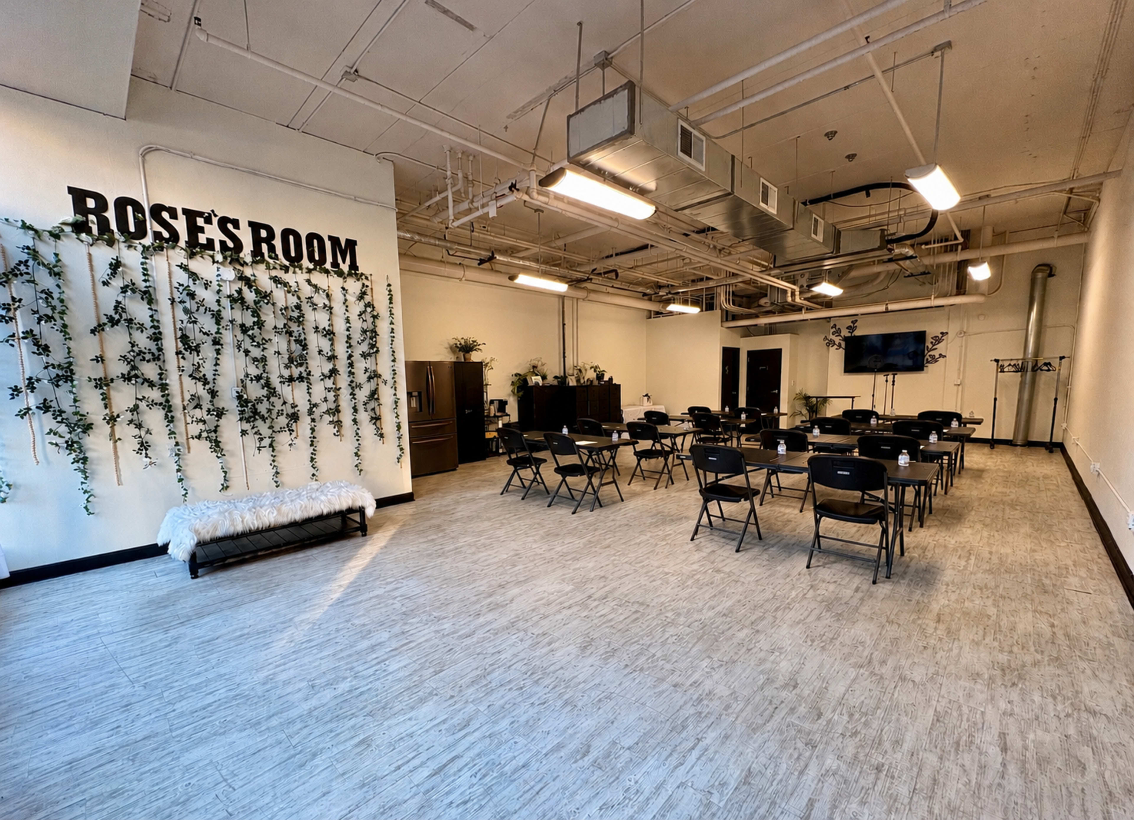 The image shows a spacious room labeled "Rose's Room," featuring a row of black chairs arranged around several tables, with greenery on the walls and a television mounted for presentations.