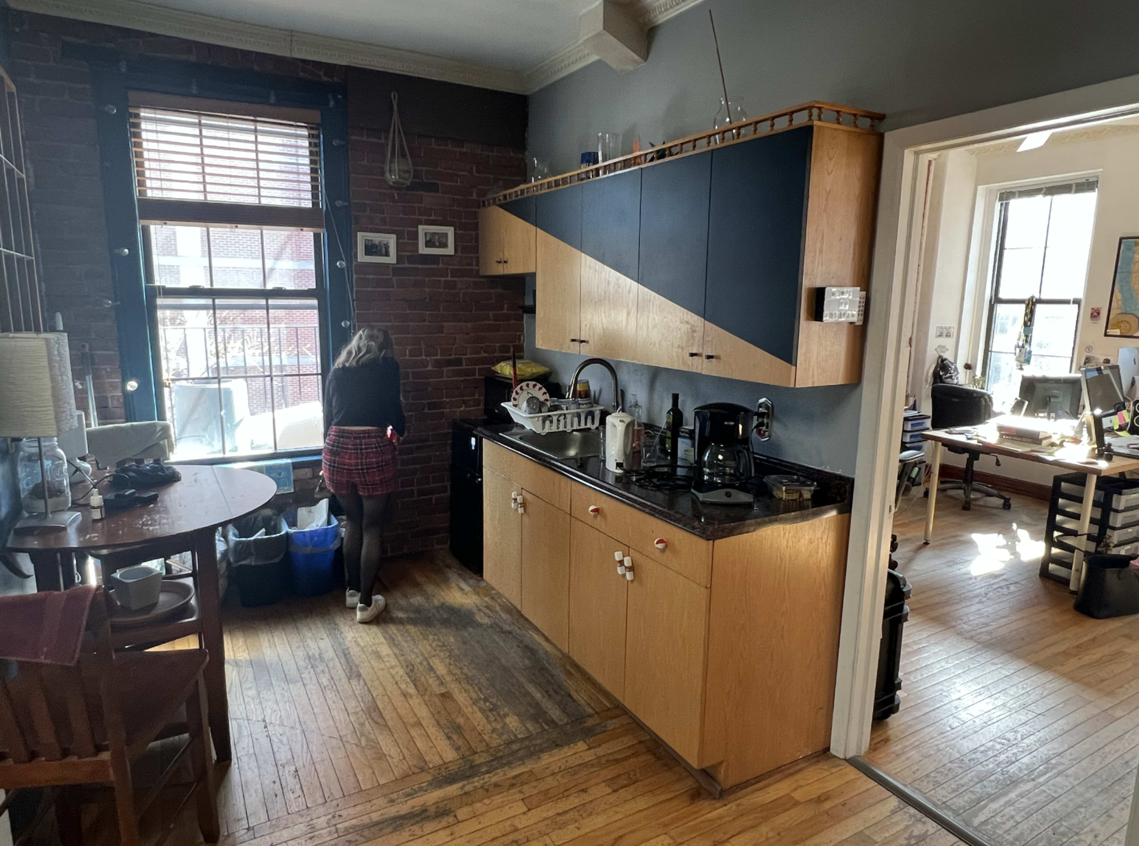 A kitchen with wooden cabinets and a black countertop is seen, featuring a person organizing items near a window, while a workspace is visible in an adjacent room.