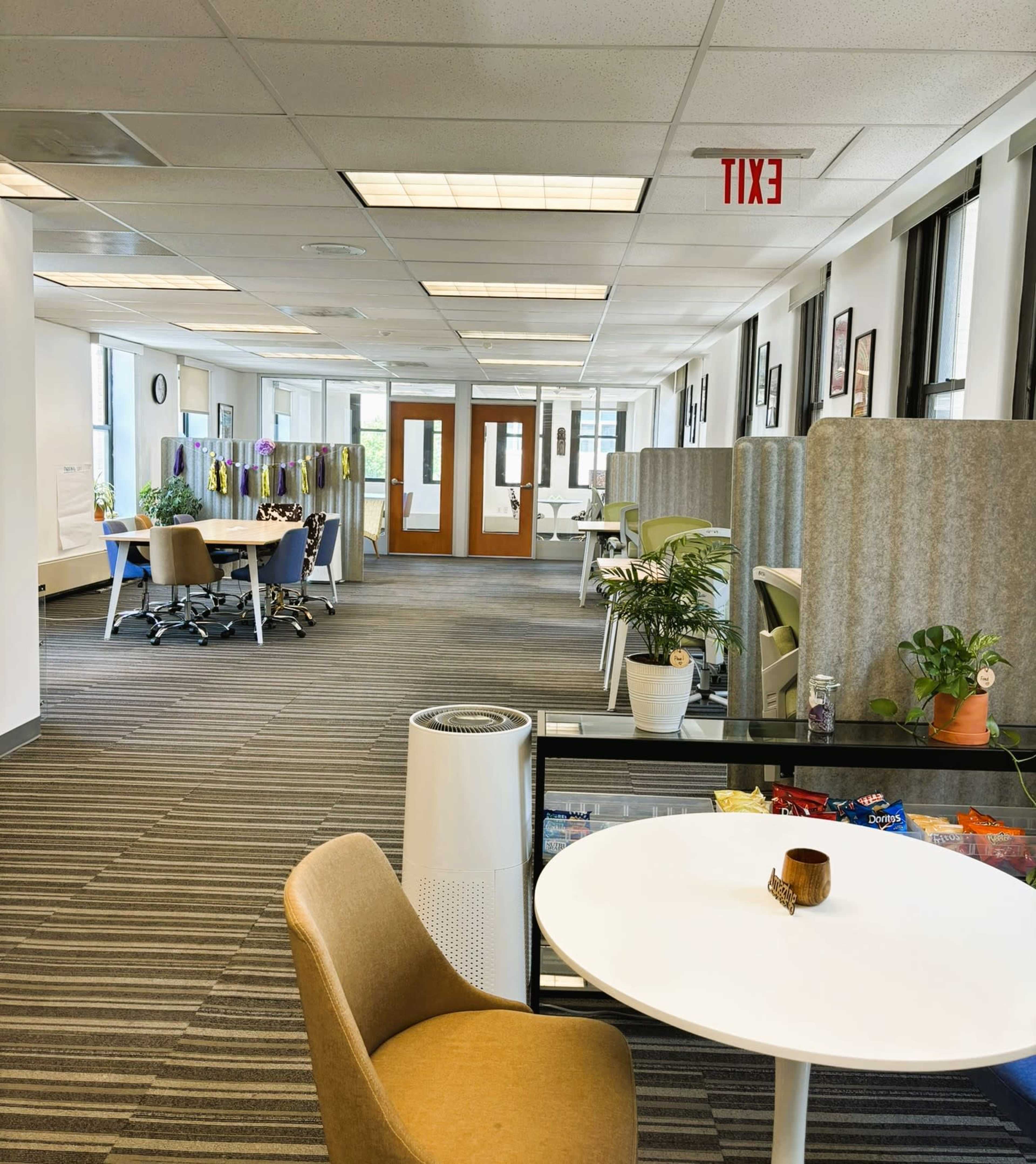 A modern office space featuring open cubicles, a communal table with snacks, and large windows providing natural light.