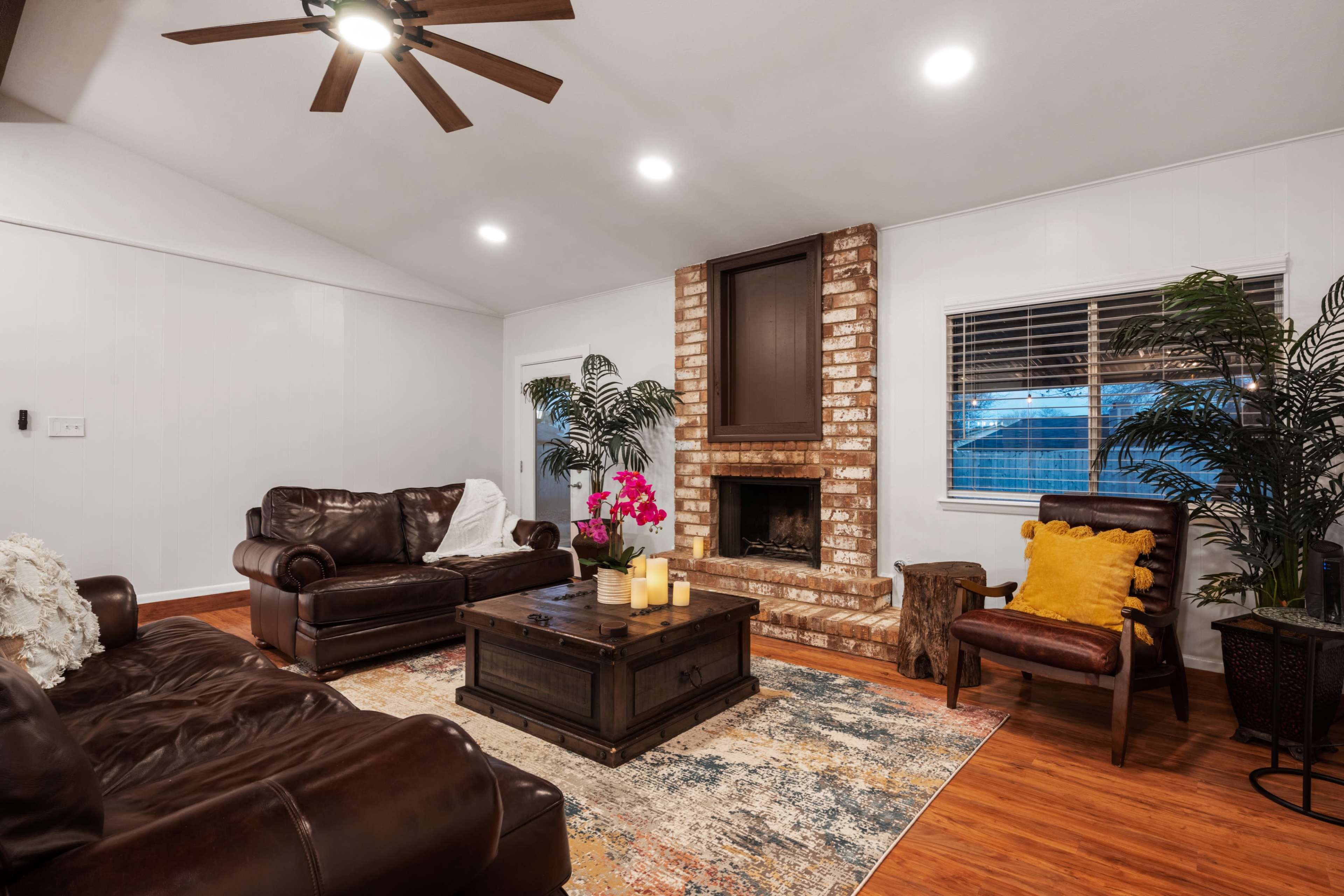 The living room features a brick fireplace, two leather sofas, and a wooden coffee table surrounded by plants and decorative elements.