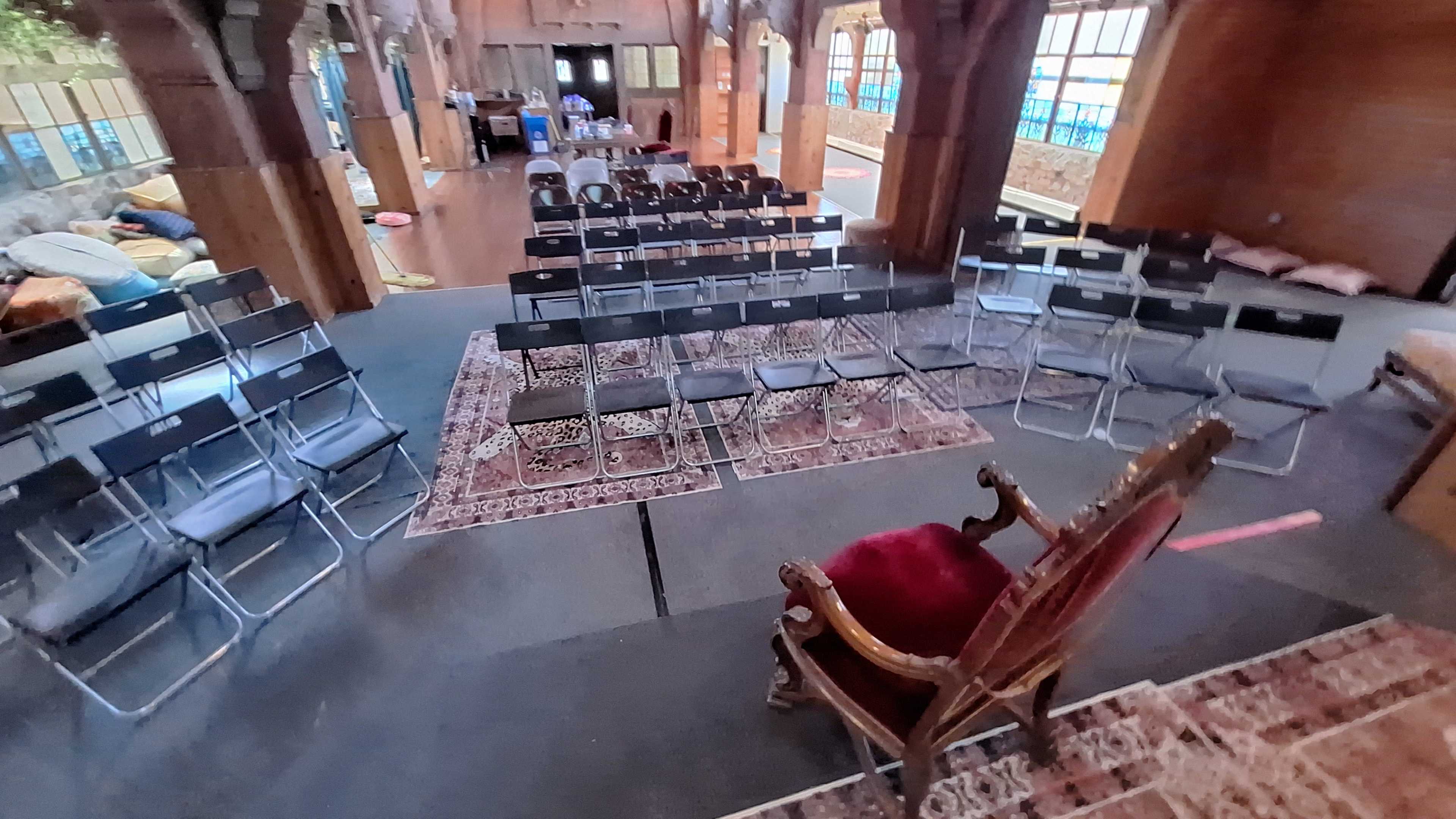 The image shows a spacious room arranged with rows of folding chairs facing an ornate wooden chair at the front, surrounded by cozy seating areas.