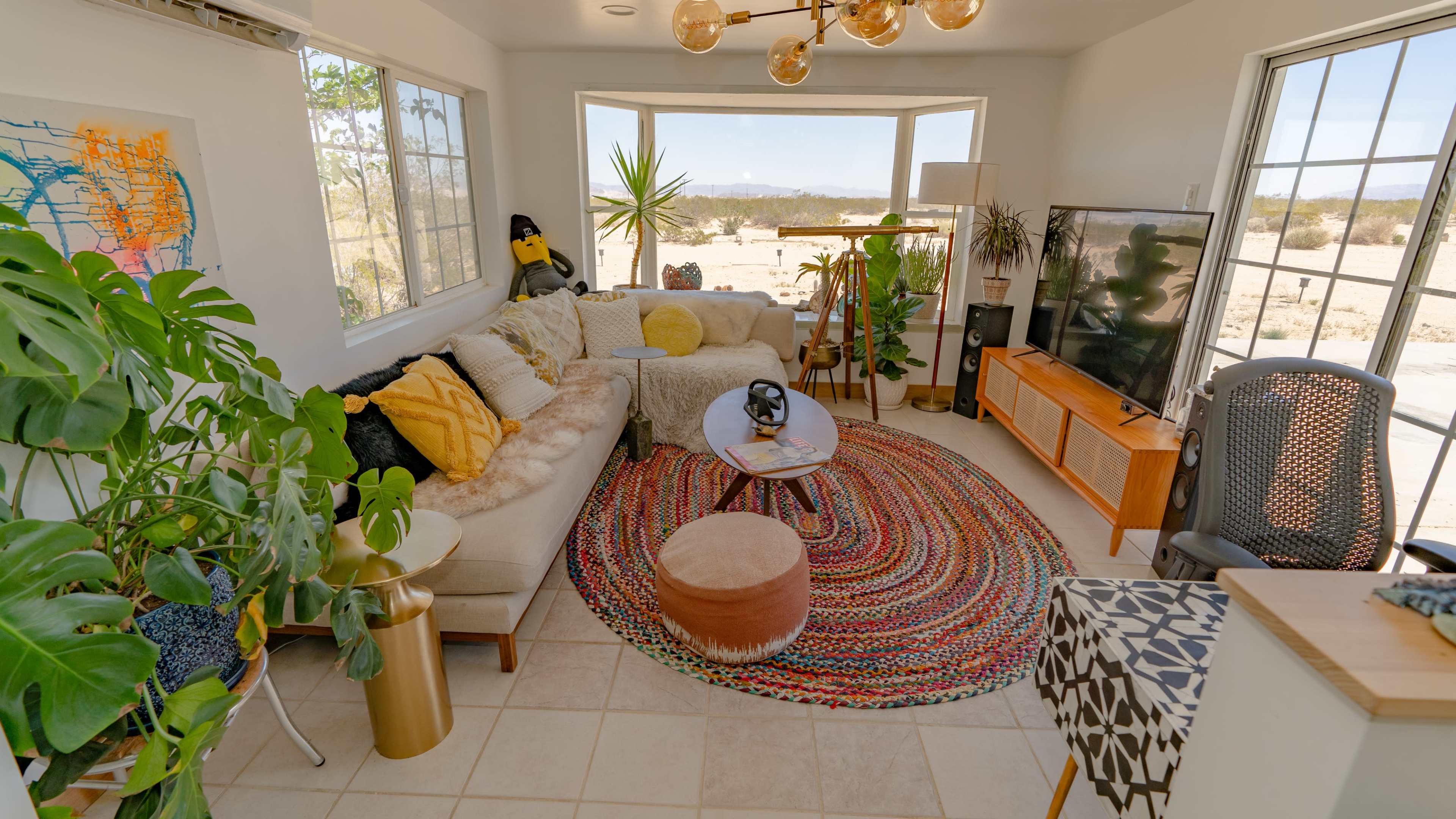 A brightly lit living room features a sectional sofa, a round coffee table, and large windows overlooking a desert landscape, surrounded by various plants and decorative furnishings.
